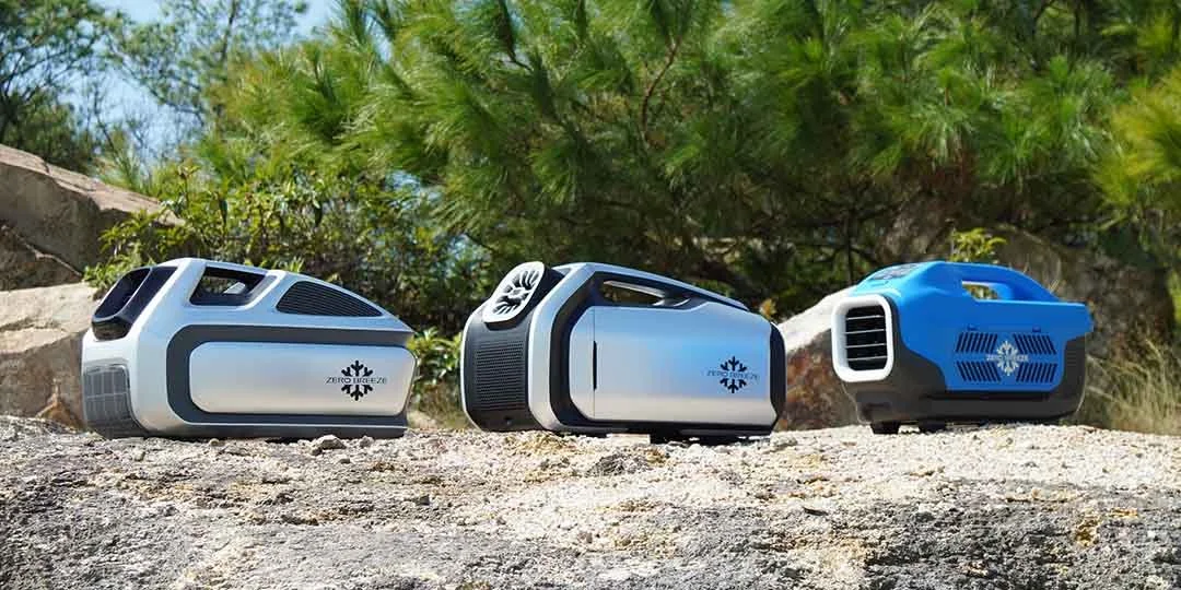 Three portable mini refrigerators or coolers placed on a rocky surface with green trees and blue sky in the background.