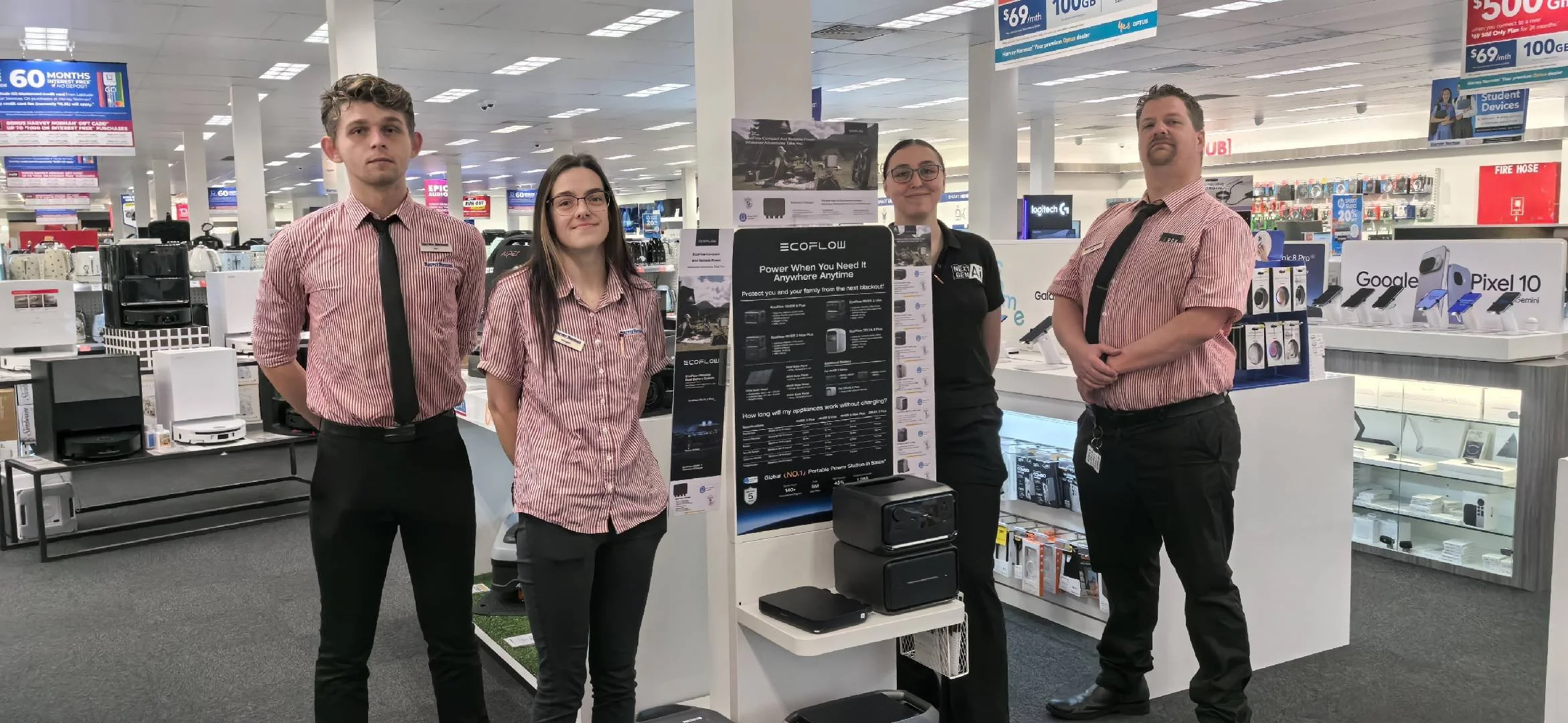 Four employees standing behind display shelves of electronics in a retail store, with advertisements and product signs visible in the background.