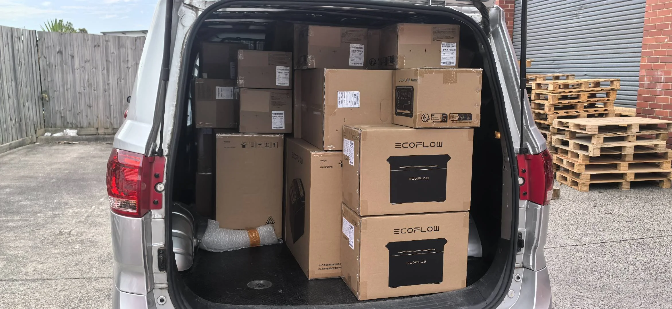 The back of a silver van filled with boxed appliances, with stacks of wooden pallets outside on a concrete lot surrounded by a brick and wood fence.