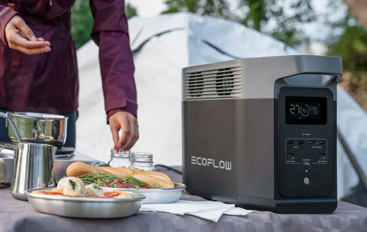 Outdoor scene with a portable ECOFLOW generator on a table, food items including pizza, bread, and herbs, and a person preparing a jar in the background.