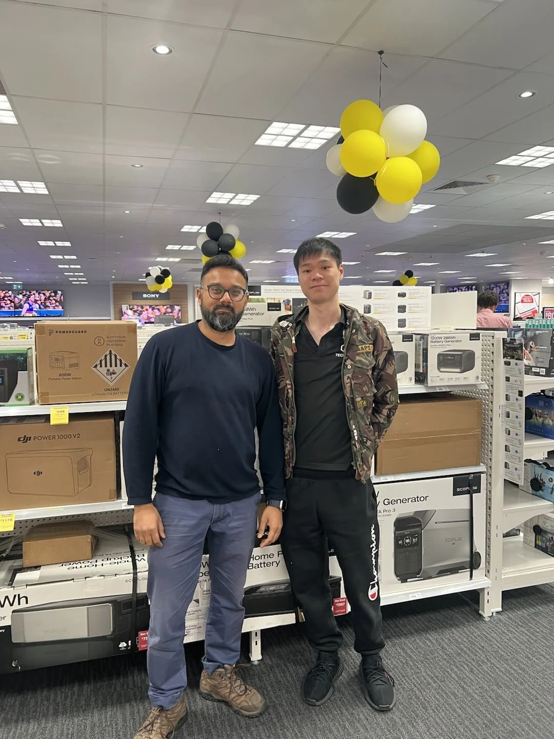 Two men standing together in an electronics store, surrounded by shelves of power generators and other electronic equipment, with black, white, and yellow balloons hanging from the ceiling.