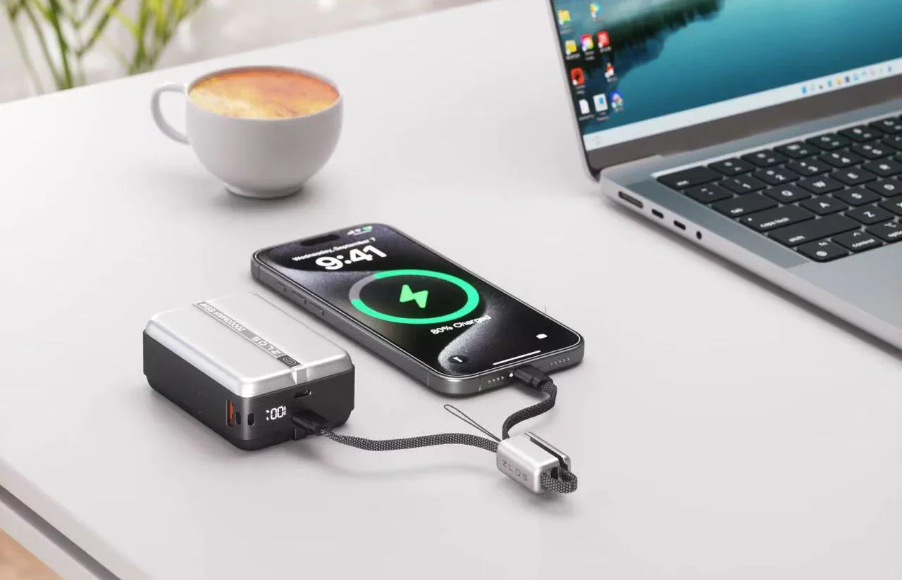 A laptop, a smartphone charging via a portable power bank, a cup of coffee, and a potted plant on a white desk.