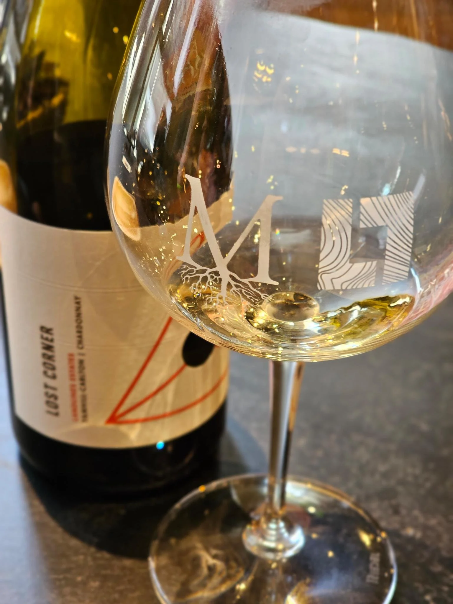 A glass of white wine with the Montinore/Landlines logo, with a bottle of Montinore Lost Corner Chardonnay in the background.