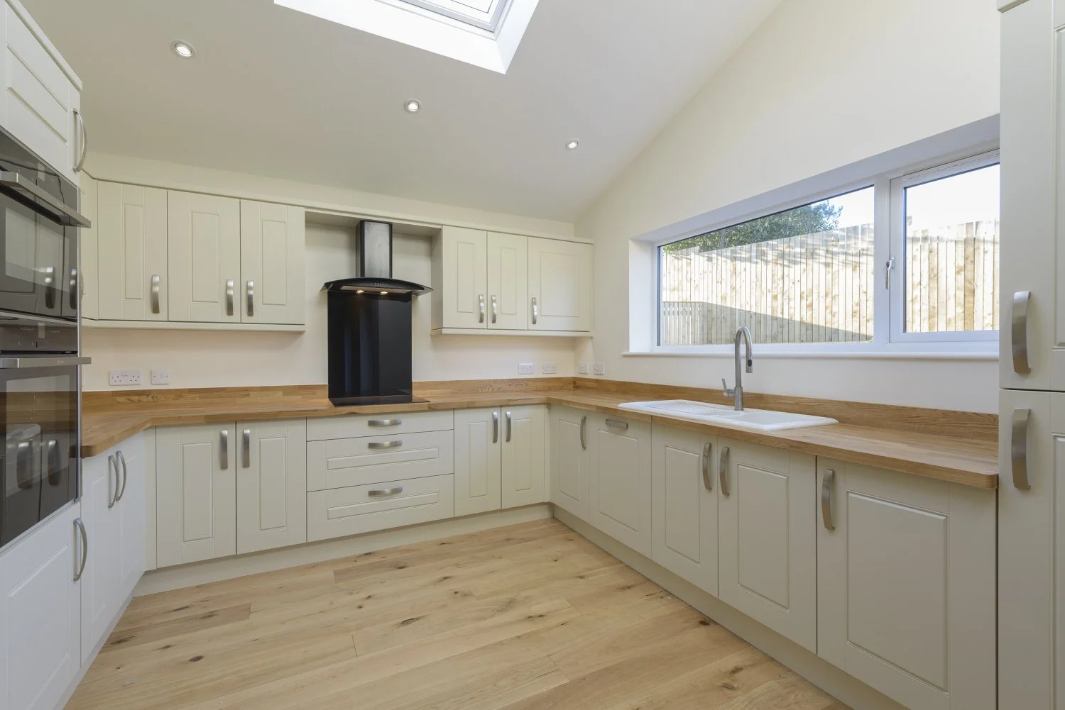 Empty modern kitchen with white cabinets, wooden countertops, a large window, and a skylight, with appliances and a black range hood.