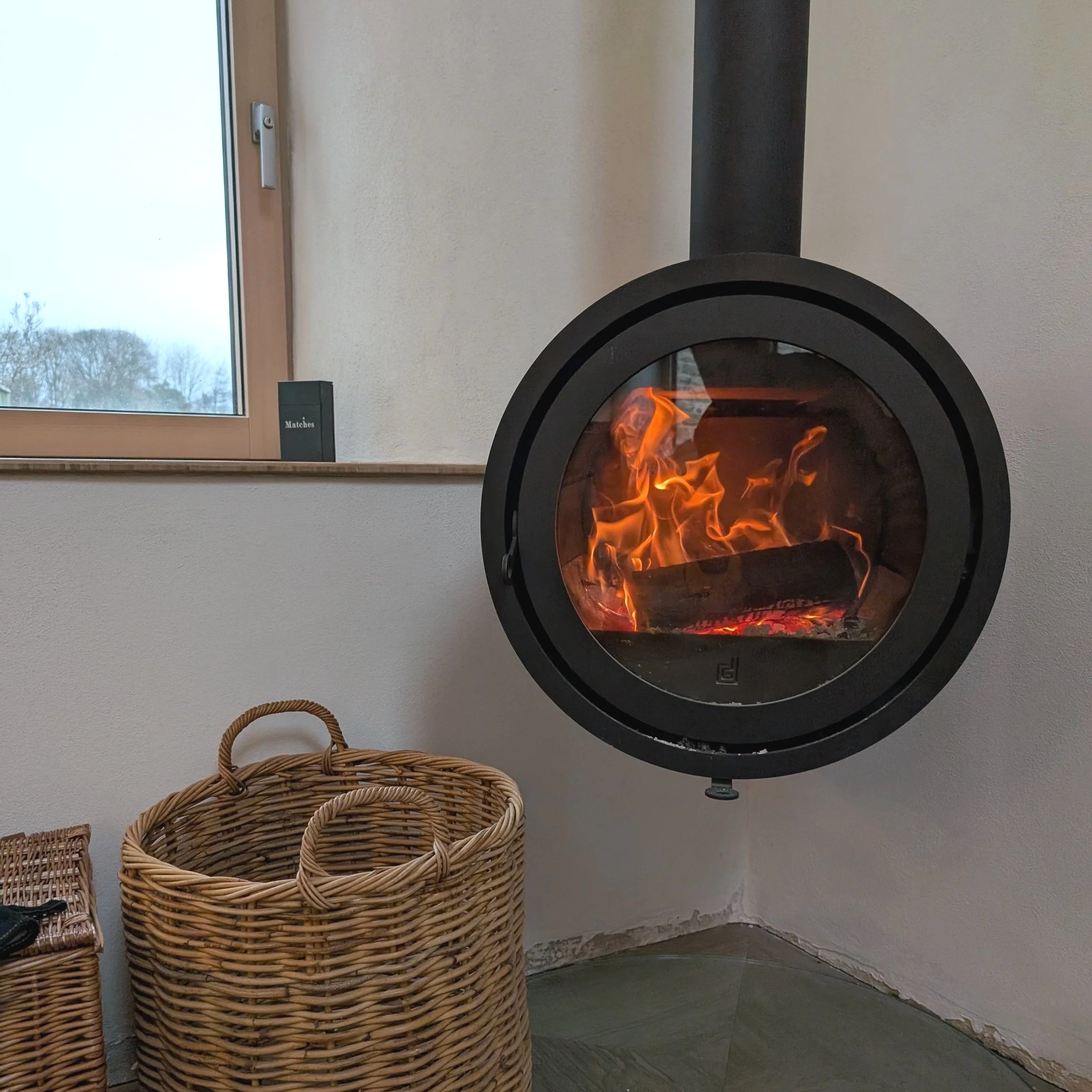 A sleek, black circular hanging woodburning stove suspended from a double-height ceiling, serving as the central architectural focal point in the vaulted living room of Owlet House