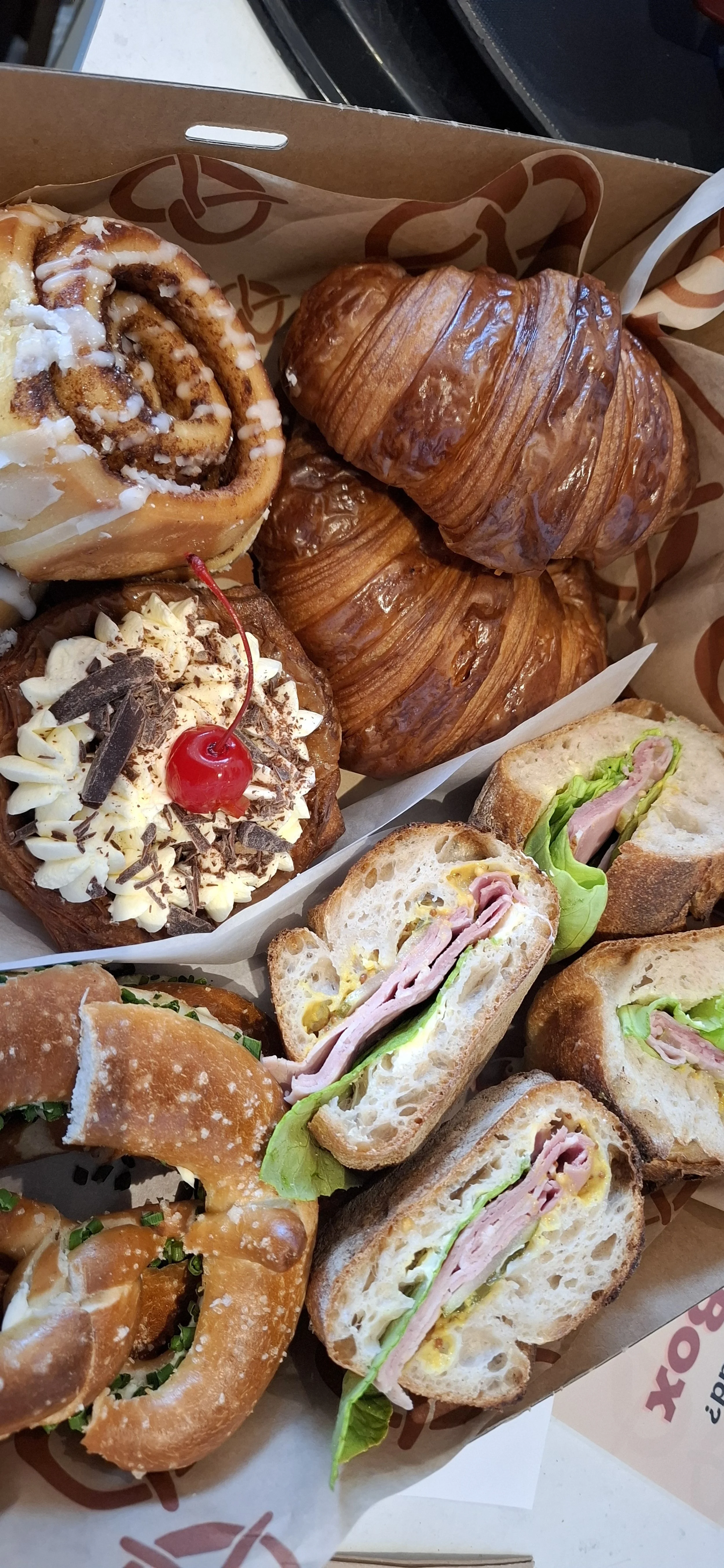 A bakery box containing assorted baked goods and sandwiches, including croissants, cinnamon rolls, a Danish pastry with a cherry on top, and sandwiches with ham, cheese, lettuce, and bread.