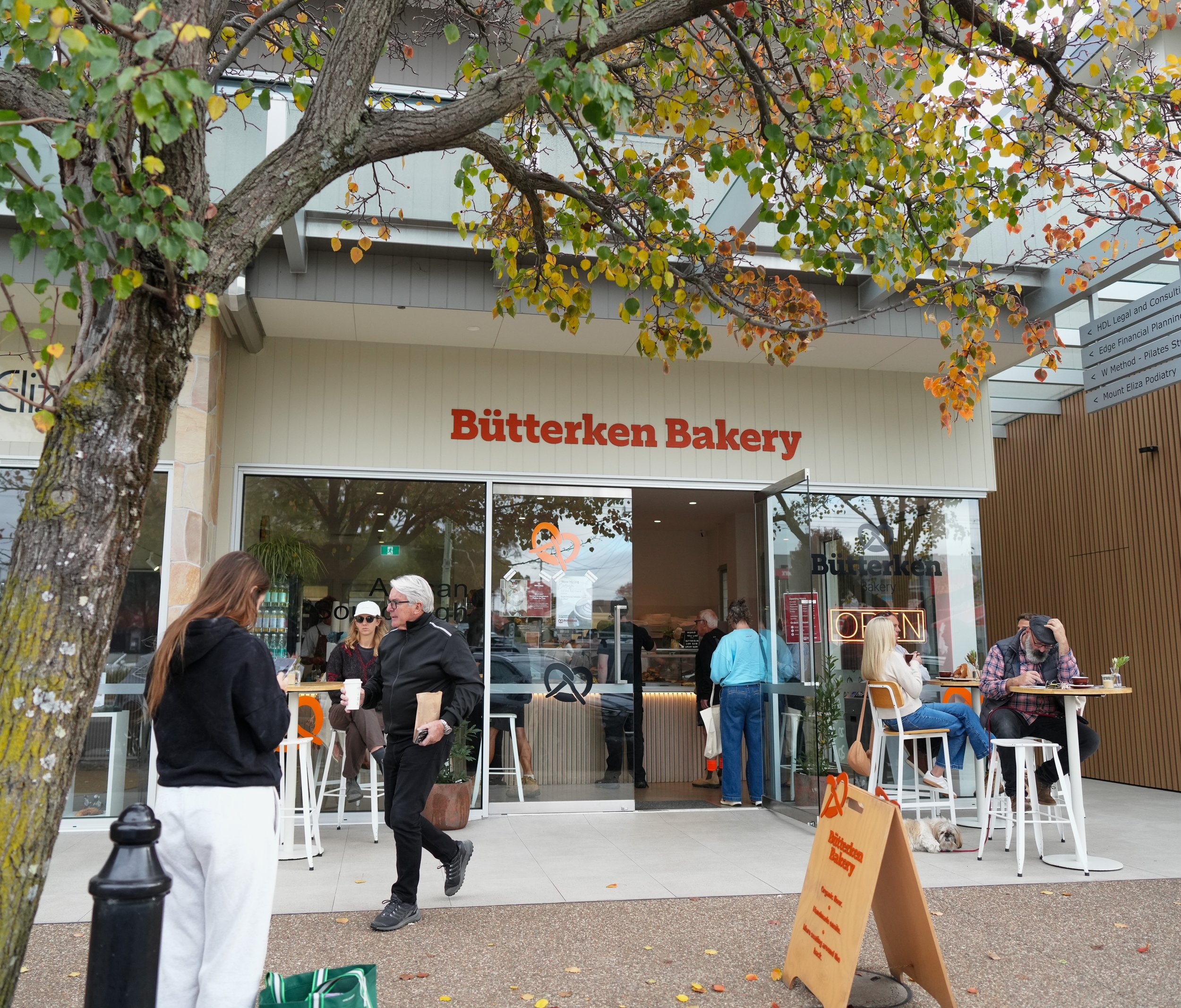 People standing and chatting outside a bakery named Bütterken Bakery, with some seated at high tables, and a dog lying next to a sign.