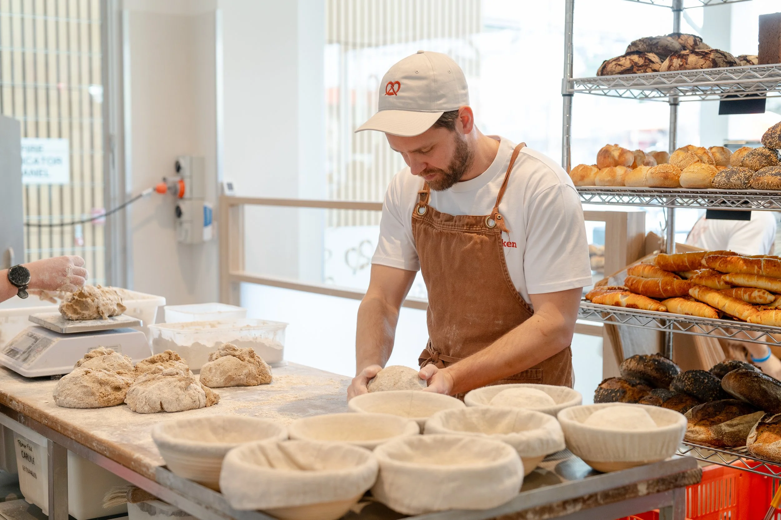 A baker wearing a white cap and apron working with dough at a bakery counter with racks of baked bread and pastries behind him.
