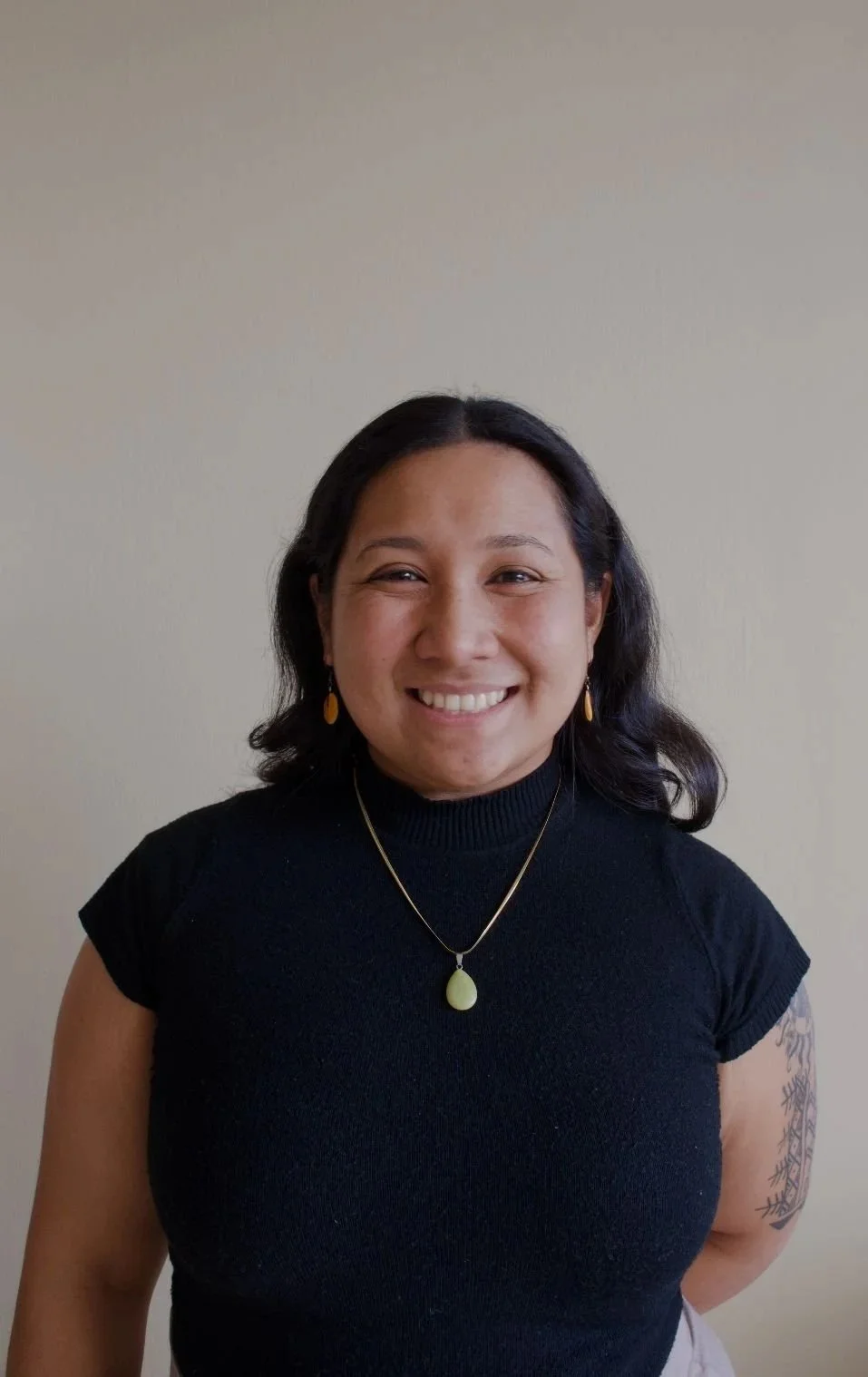 A smiling woman with dark hair wearing a black shirt, earrings, and a gold necklace with a large green pendant, standing against a plain beige background.