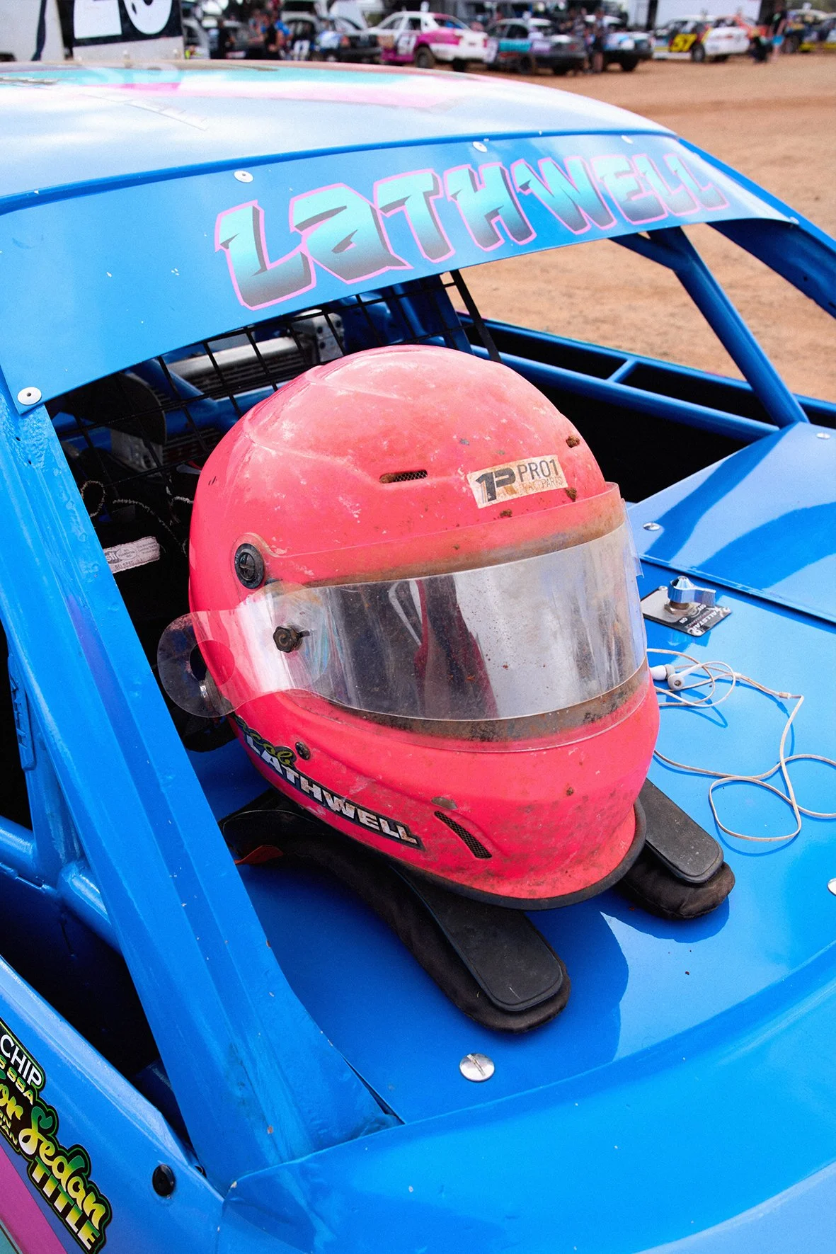 A pink racing helmet with a clear visor and black detail, placed on the dashboard of a blue race car at a dirt track event, with cars and spectators visible in the background.