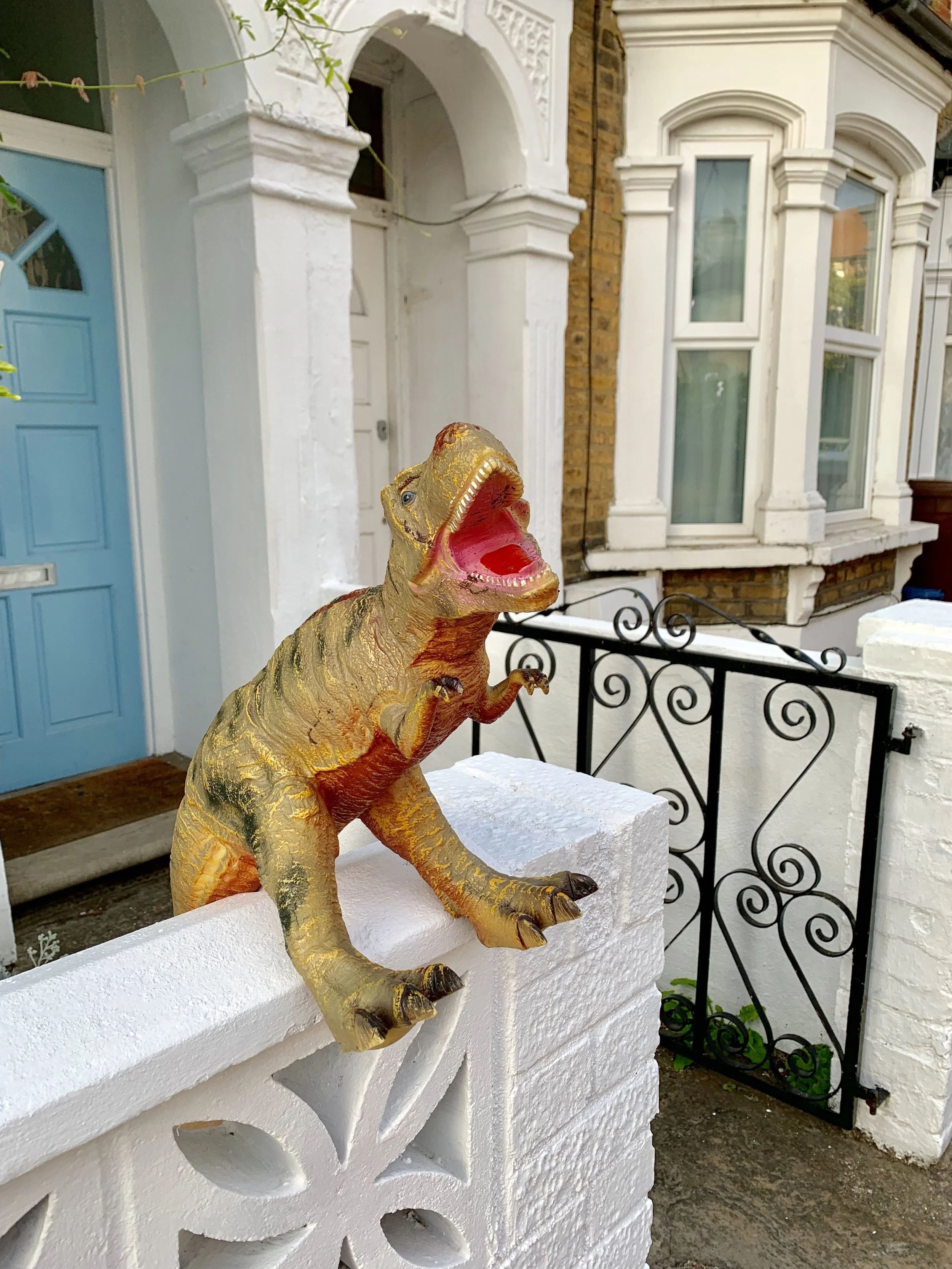 A humorous plastic dinosaur with an open mouth, painted in gold and red, sitting on a white brick wall outside a house.