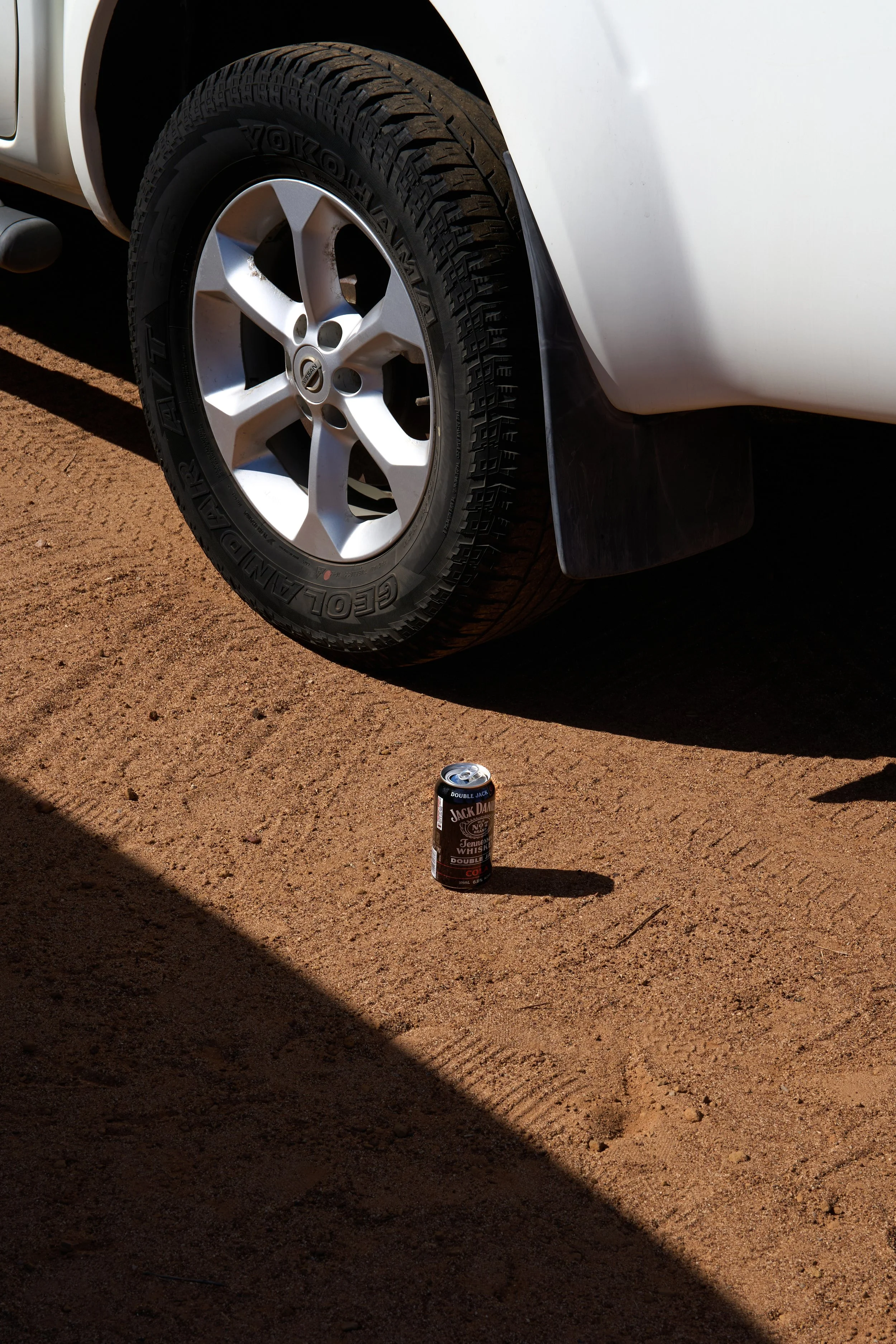 A close-up of a white vehicle's front tire and silver alloy rim on a dusty ground, with a can of Jack Daniel's whiskey on the ground below the tire, casting a shadow.