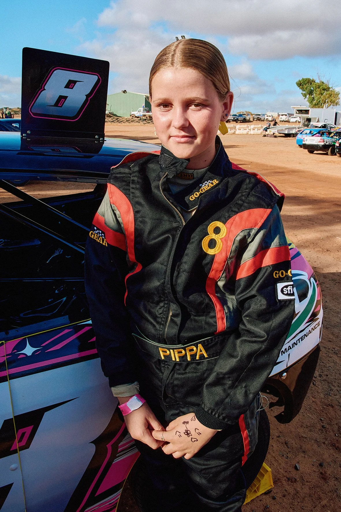 A young female race car driver with a racing suit standing next to a race car in a dirt race track on a cloudy day.