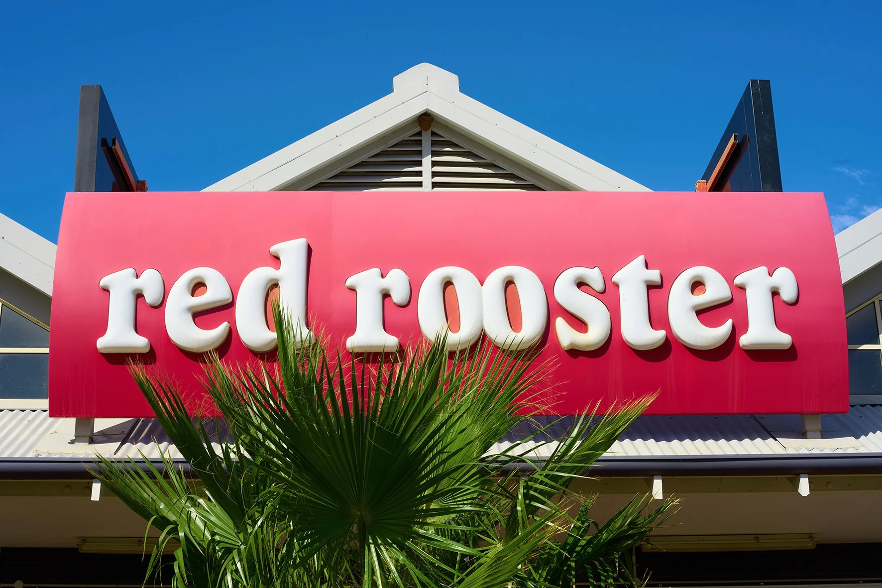 Sign for Red Rooster with white 3D letters on a pink background, topped by a white building with a gable roof and blue sky, with green palm leaves in front.