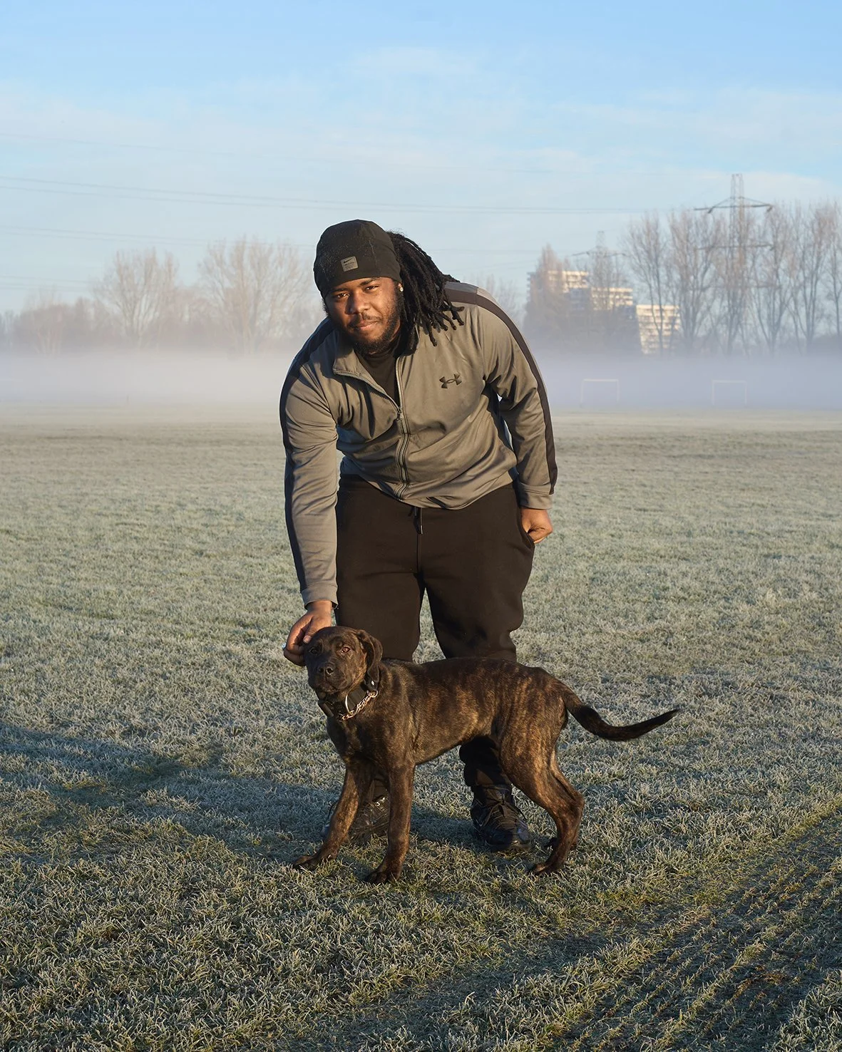 A man with dreadlocks wearing a gray jacket and black pants is standing on a foggy field with a brindle puppy.