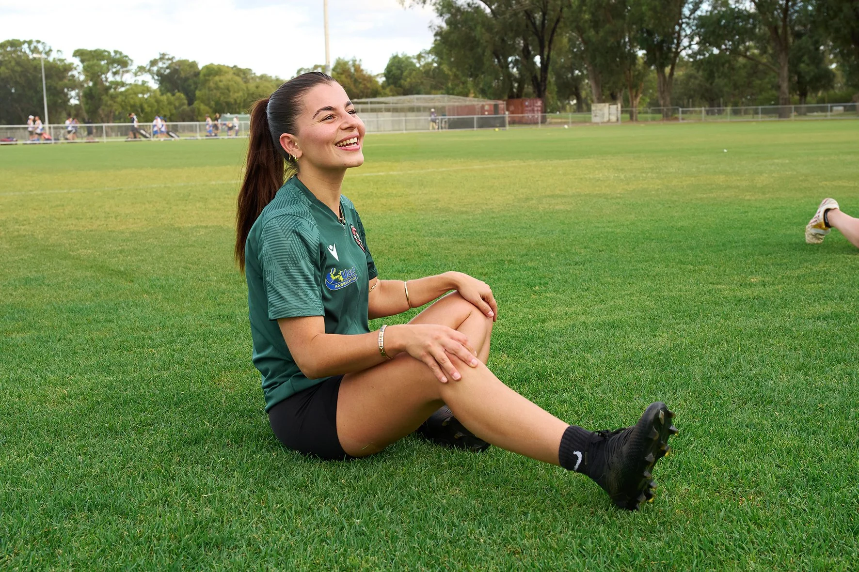 A woman sitting on the grass field of a soccer field, smiling, wearing a dark green sports jersey, black shorts, and black soccer cleats.