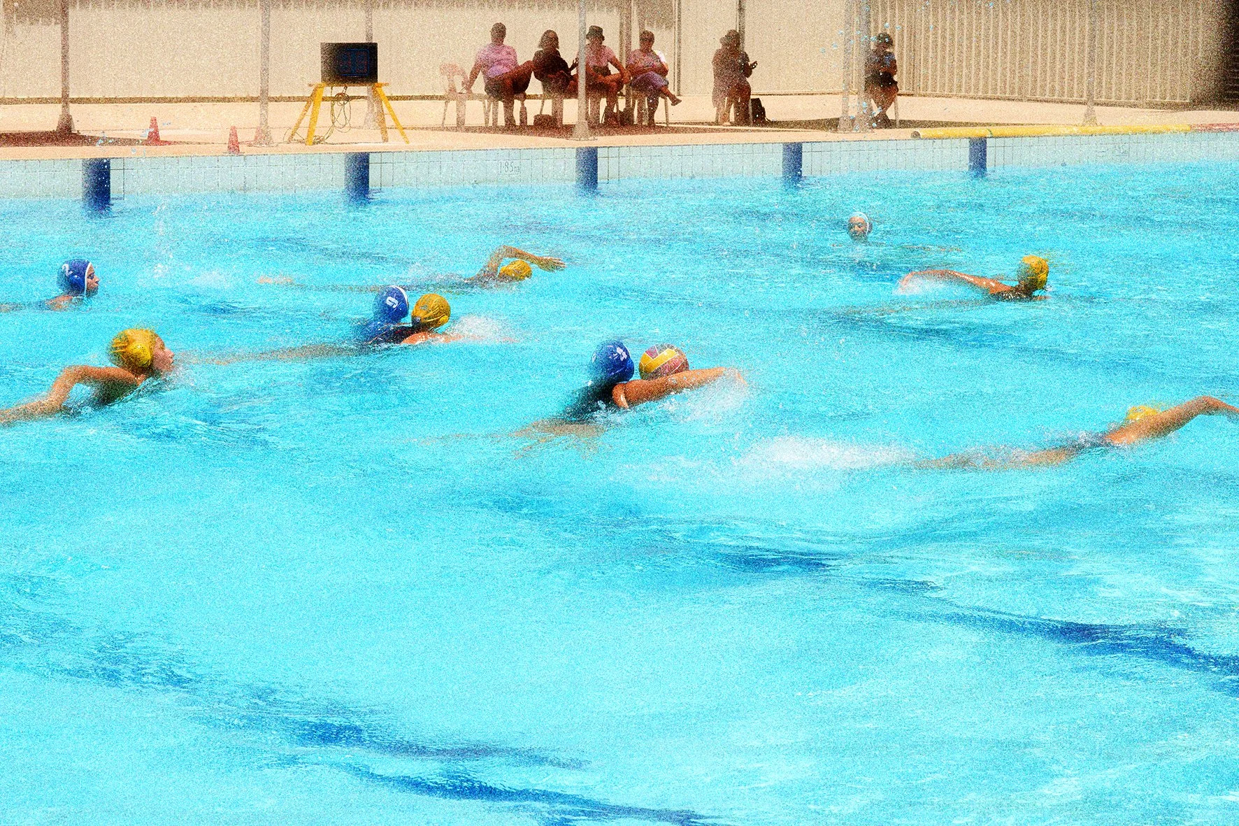 A group of people playing water polo in an indoor swimming pool, with some players in blue caps and others in yellow caps, and spectators sitting on chairs at the poolside.