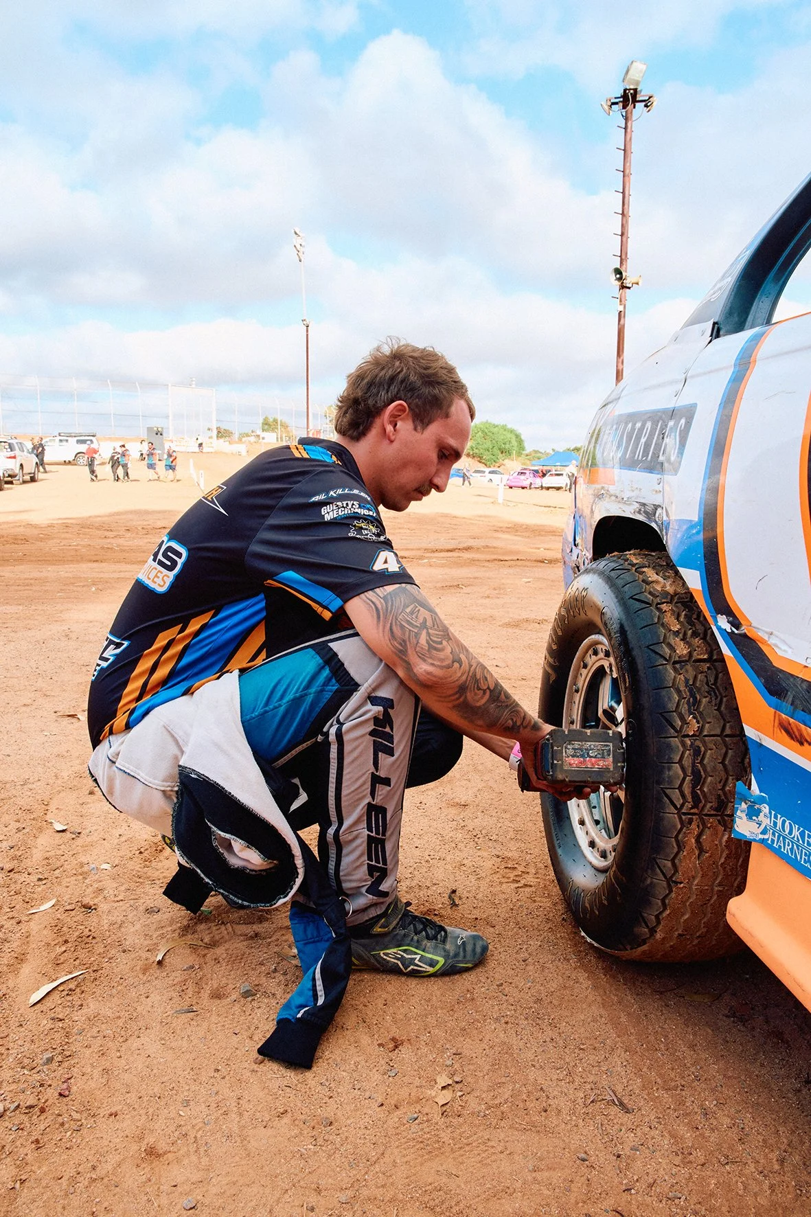 A man with a racing uniform and tattoos changing a tire on a race car at an outdoor dirt track.