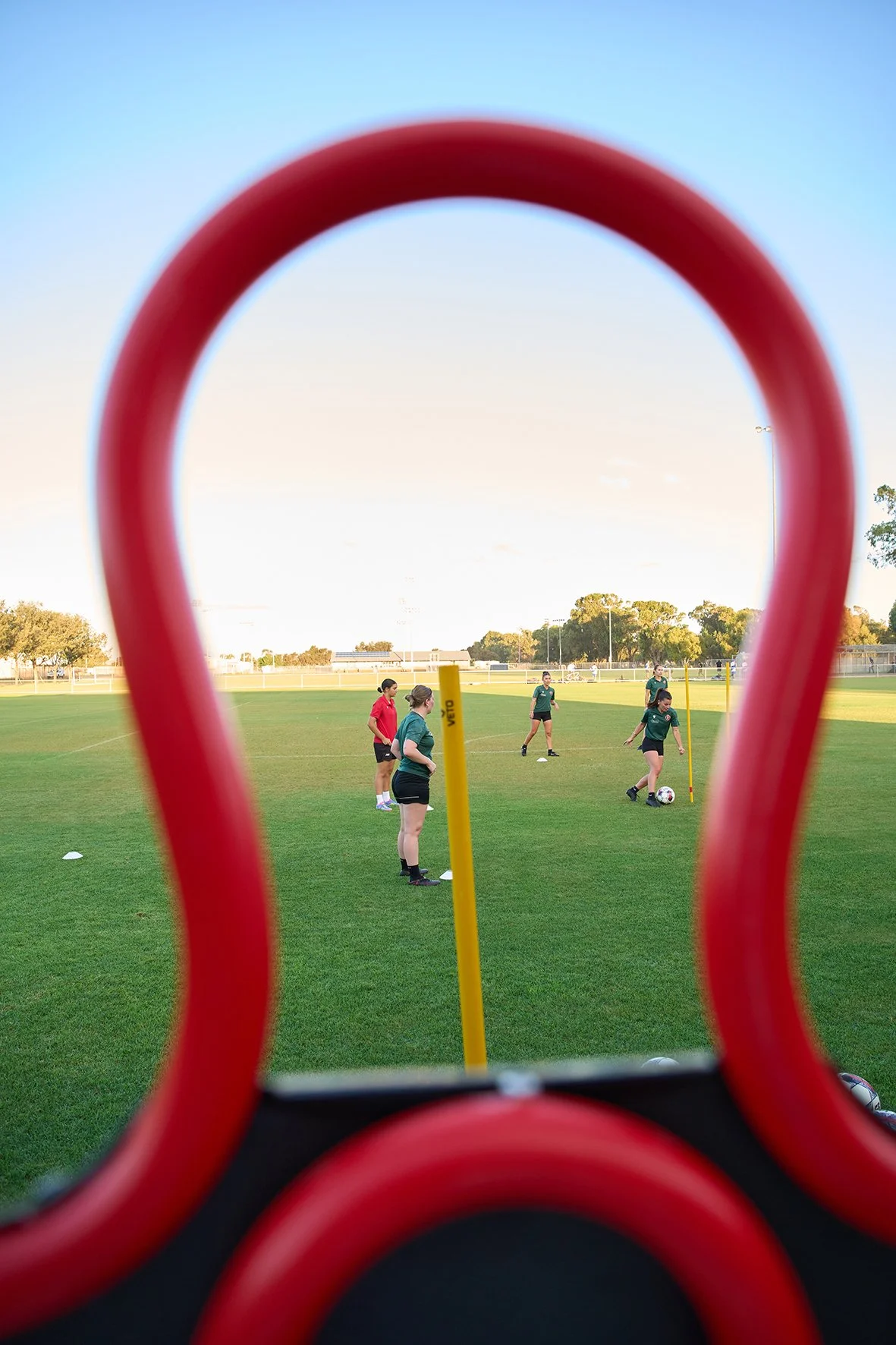 Soccer players from LaFiamma FC practicing on a field, seen through a red training device, with four females in green uniforms and one in red, some with soccer balls and cones, during daytime.