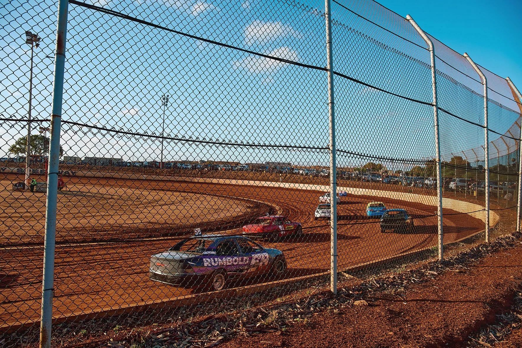 Race cars on a dirt track behind a chain-link fence during a race in the daytime.