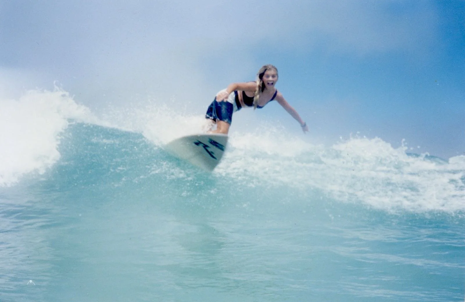 A girl surfing on a wave in the ocean, wearing a black top and blue shorts, with an excited expression on her face.
