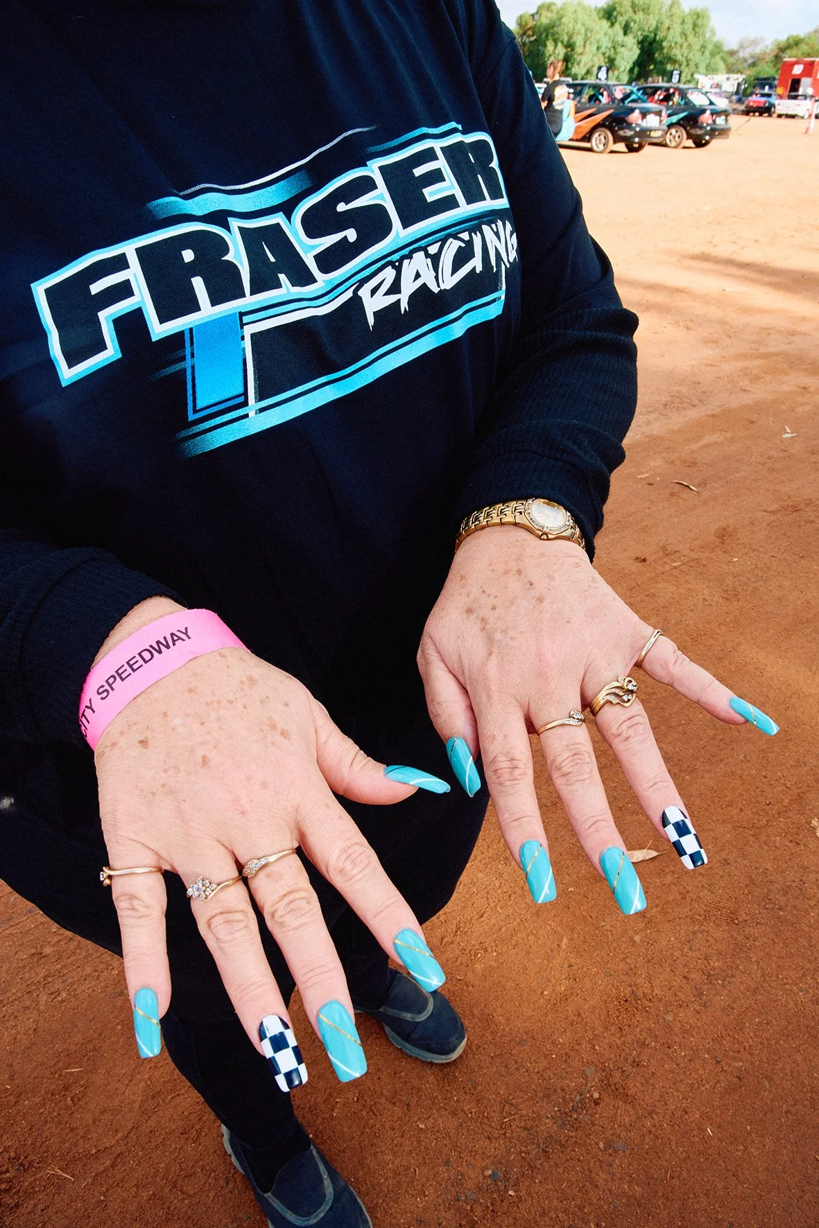 Person wearing a Frasher Racing sweatshirt, showing off their decorated blue and checkered nails, rings, and a gold wristwatch at a dirt racing event with cars and trees in the background.