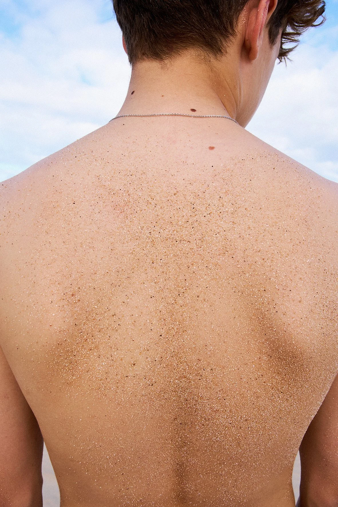 Back of a person with sand on their shoulders and neck, wearing a thin necklace, outdoors with a blue sky and clouds.
