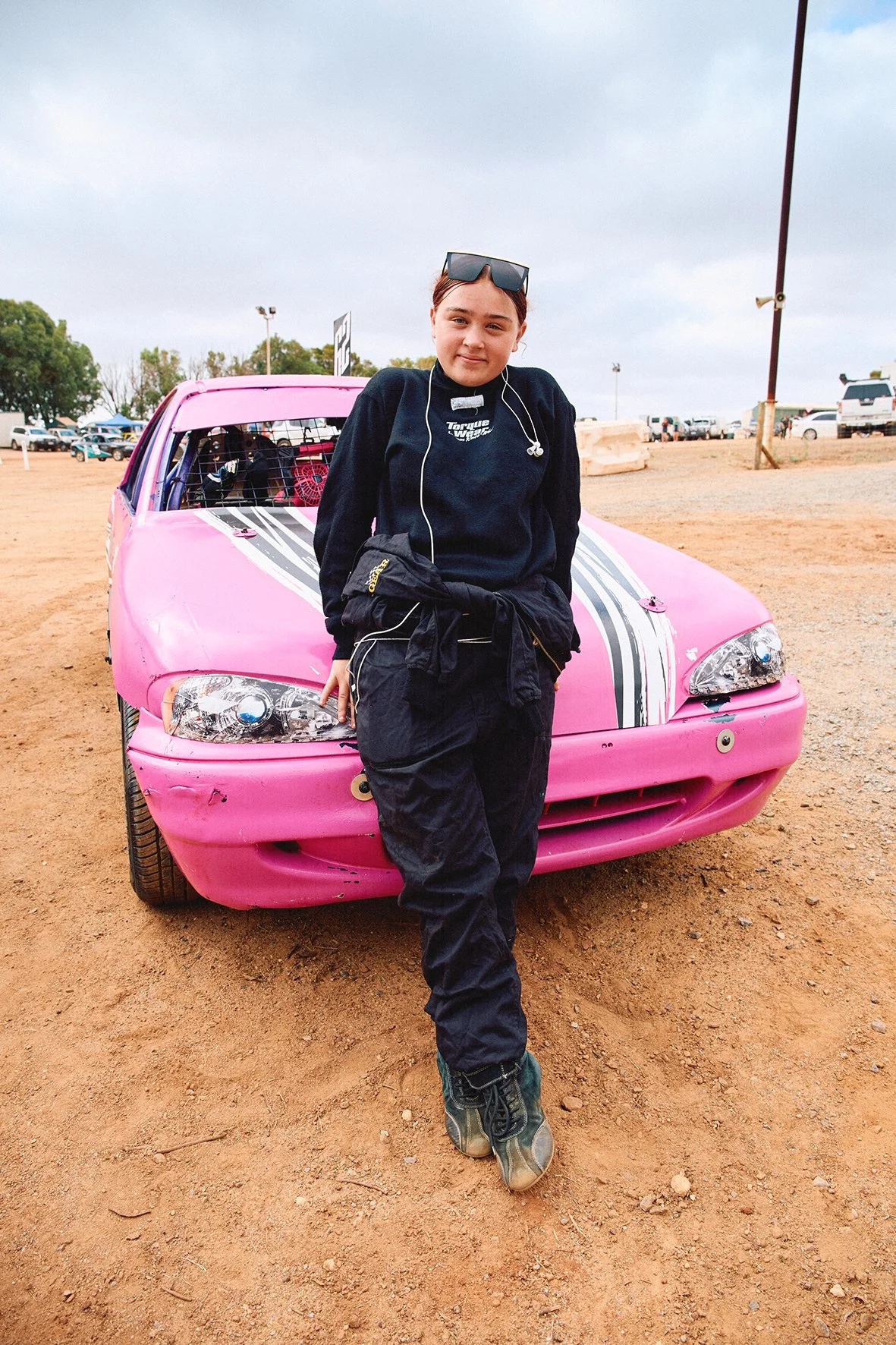 A young girl standing in front of a pink racing car at a dirt track, wearing racing gear and sunglasses.