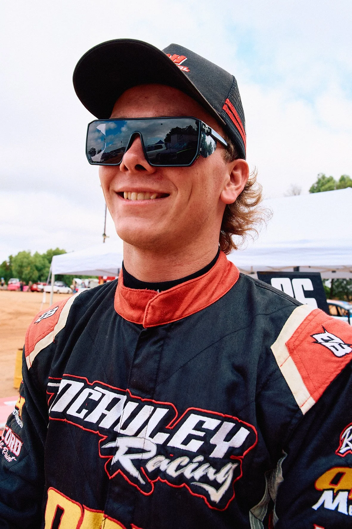 A smiling race car driver wearing a black and red racing suit with sponsor logos, large reflective sunglasses, and a black cap with red accents. Background shows a race track and tents.