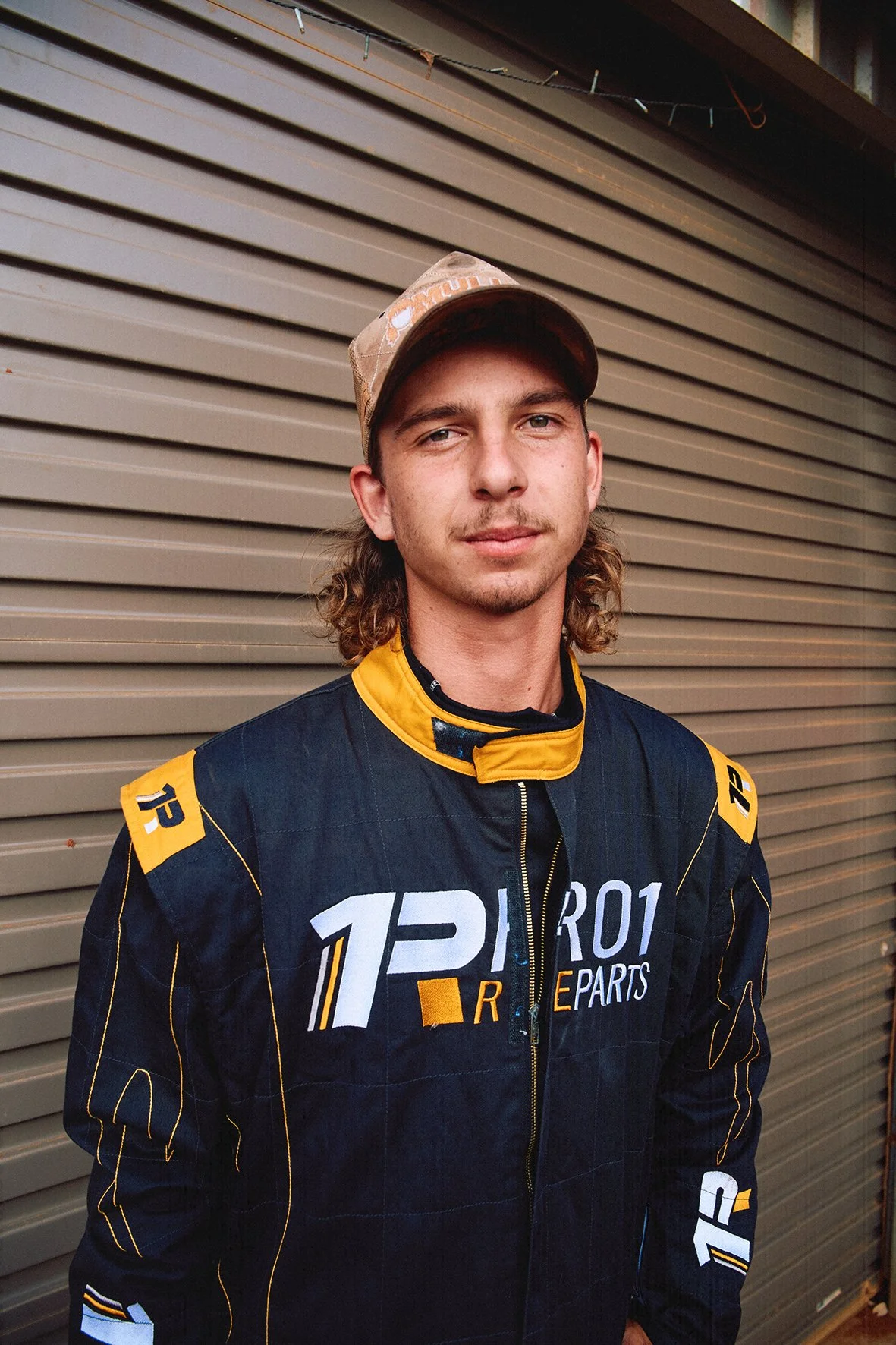 A young man in a racing suit and baseball cap standing in front of a metal garage door.