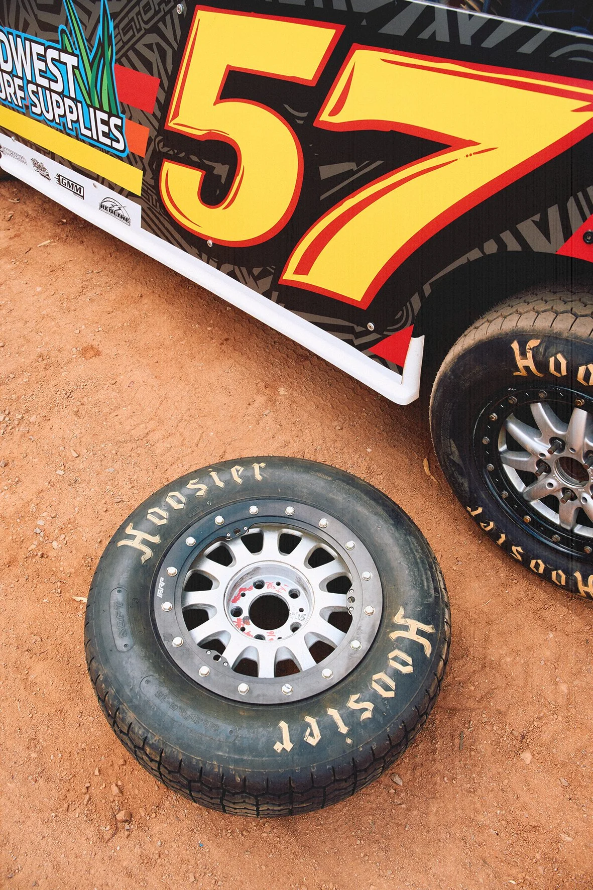 A race car on a dirt track with its tires removed. The car has a large number 57 and sponsor logos on its side, including 'Midwest Surf Supplies' and 'Hoosier' tire brand.