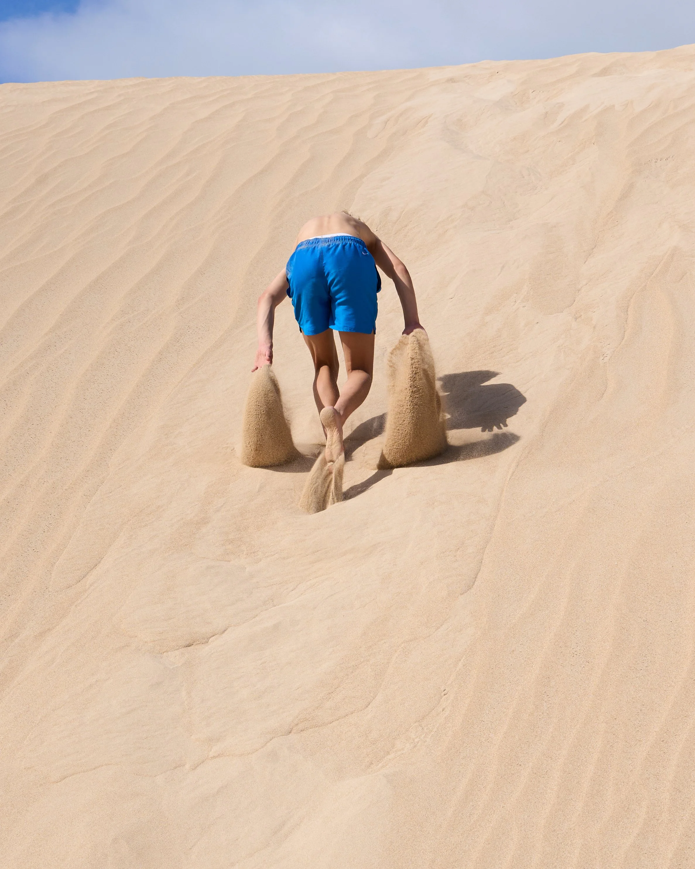 A person climbing up sand dunes on all fours, wearing blue shorts and no shirt, with sand falling from their hands.