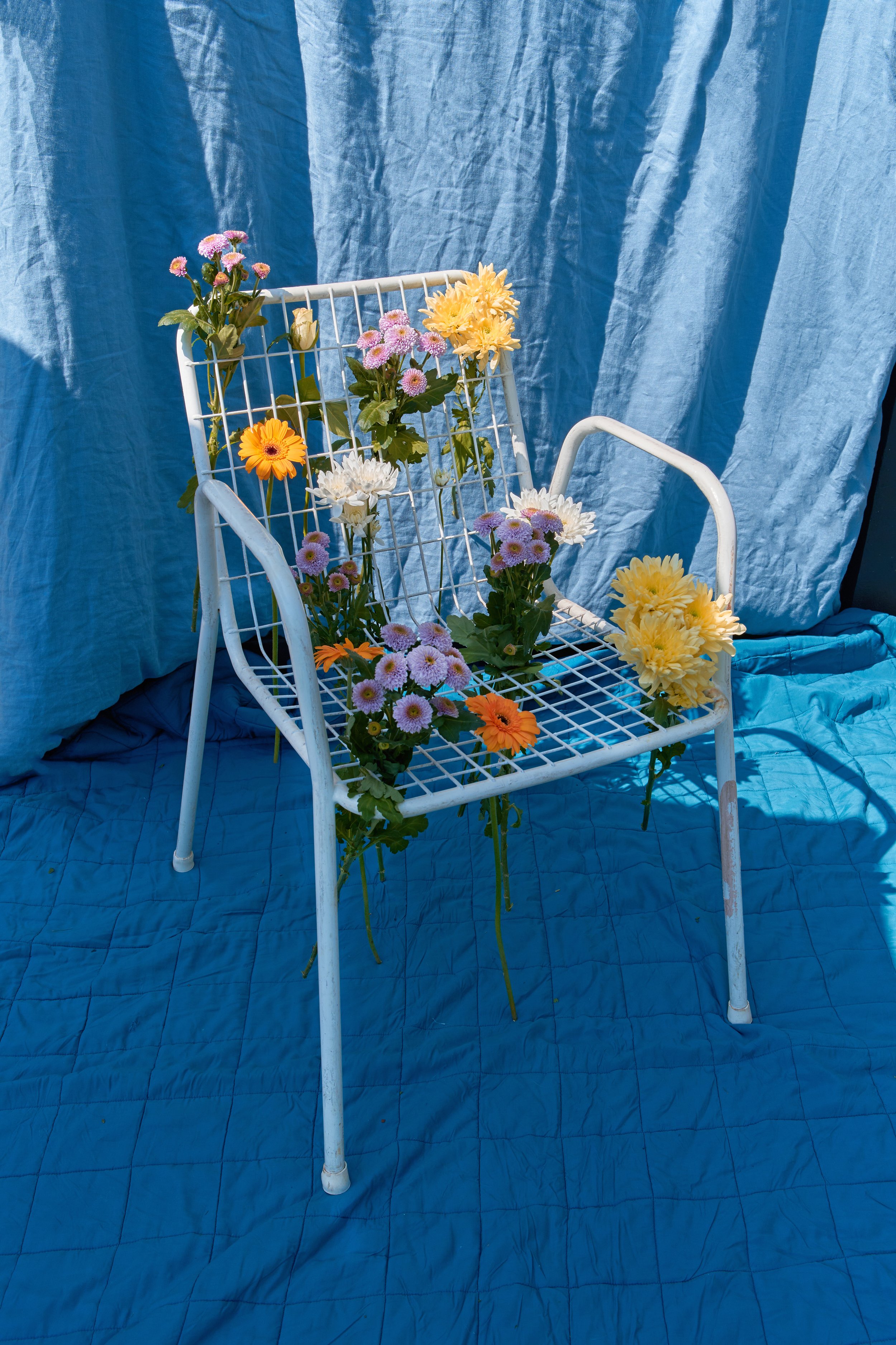 A white metal chair with blue fabric background and a light blue quilted surface, decorated with colorful flowers including yellow dahlias, white daisies, purple asters, and orange calendulas.