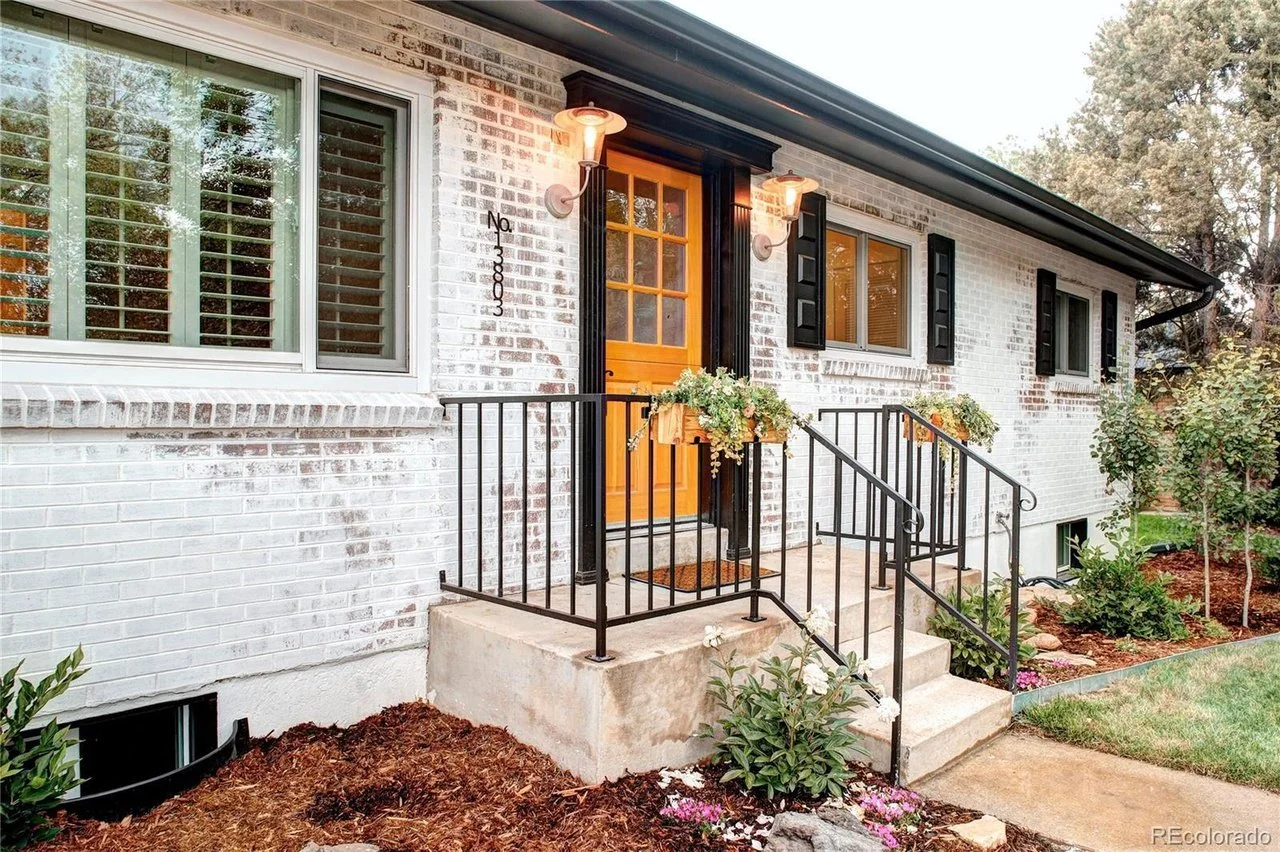 Front entrance of a white brick house with steps, black metal railings, and a wooden door with windows. There are two lantern-style lights, flowers in planters, and a landscaped garden with shrubs and plants.