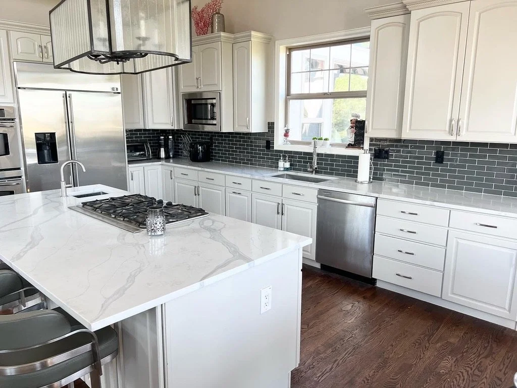 Modern kitchen with white cabinets, gray tile backsplash, marble countertops, stainless steel appliances, and an island with a sink and cooktop.