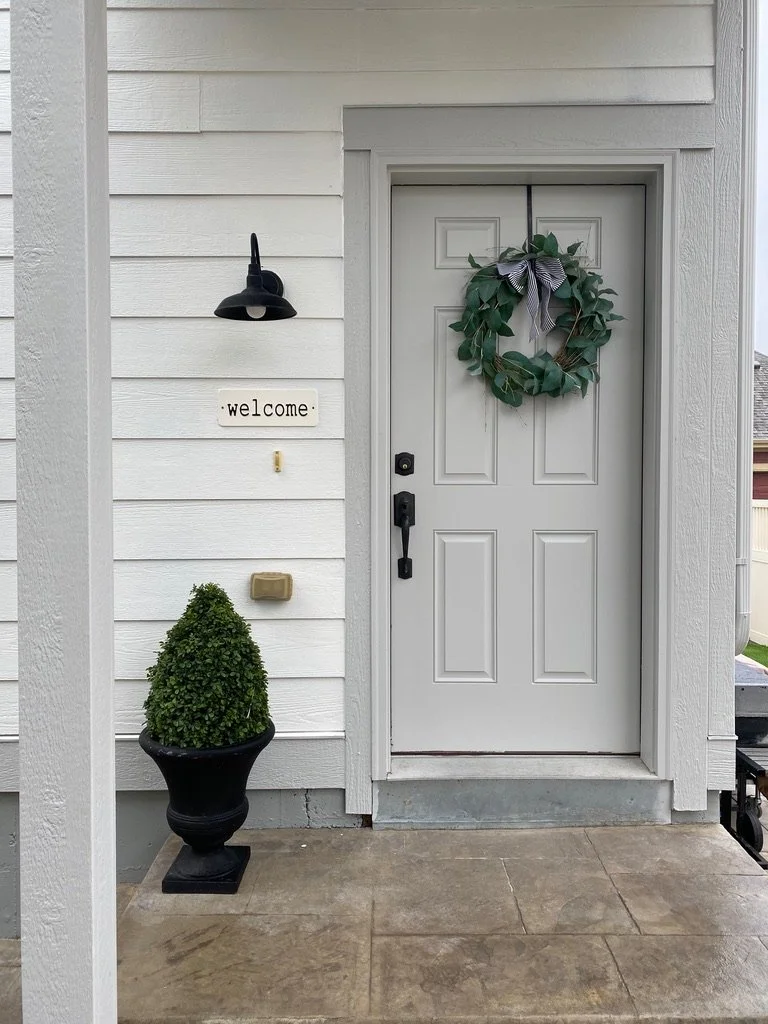 Front door with green wreath, welcome sign, and potted topiary