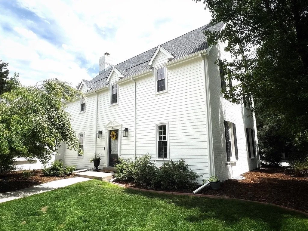 White colonial-style house with dark shutters, surrounded by trees and a well-maintained lawn.