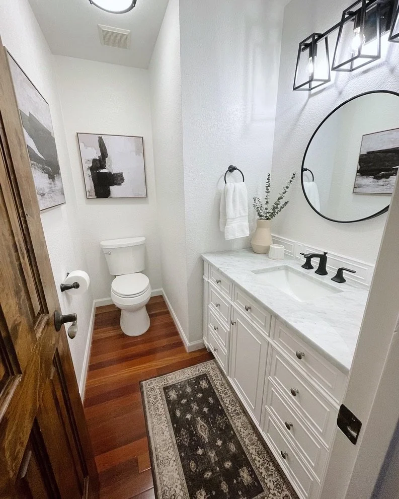 Modern bathroom with a wooden door, white walls, and hardwood floor. Features a toilet, white vanity with a marble countertop, round mirror, and decorative lighting. Includes abstract wall art, hand towel on a ring, and potted plant on the counter. A