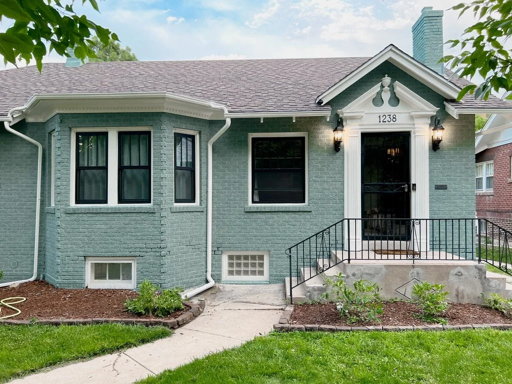 Front view of a blue-painted brick house with a small porch, black door, and black-framed windows, surrounded by a garden and lawn.
