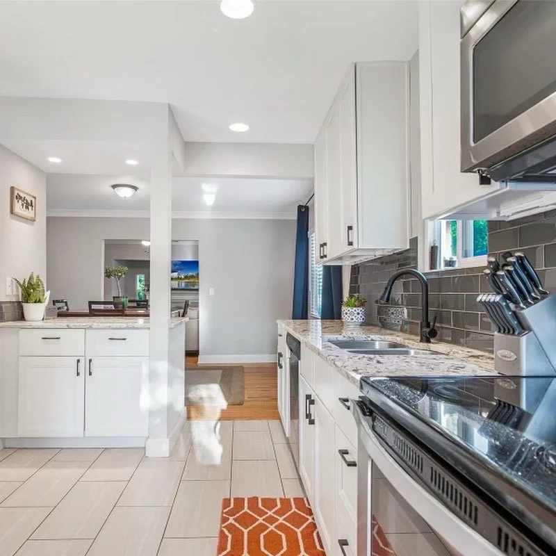 Modern kitchen with white cabinets, granite countertops, black faucet, stainless steel appliances, and a red patterned rug on tiled flooring.
