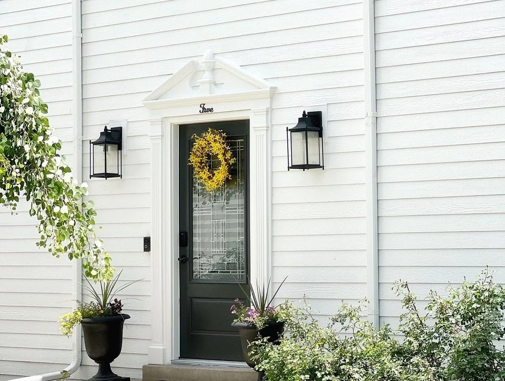 Front door with a yellow wreath, white siding, black lanterns, planter with greenery, and plants around the entrance.