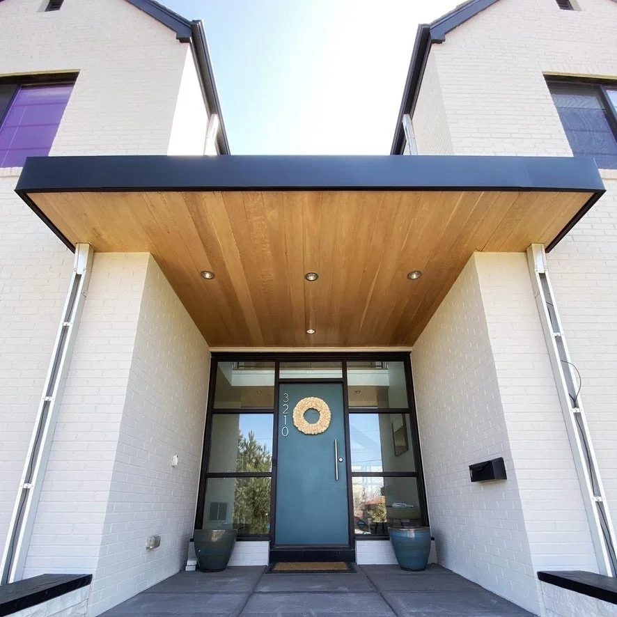 Modern home entrance with teal door, wood overhang, brick walls, and decor.