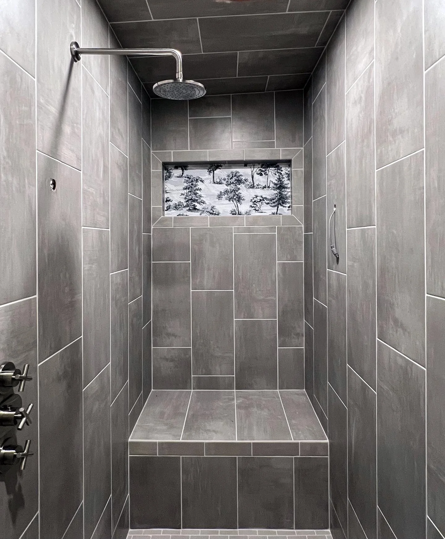 Modern gray-tiled shower with a built-in bench and a small window, featuring a rain shower head and wall-mounted controls.