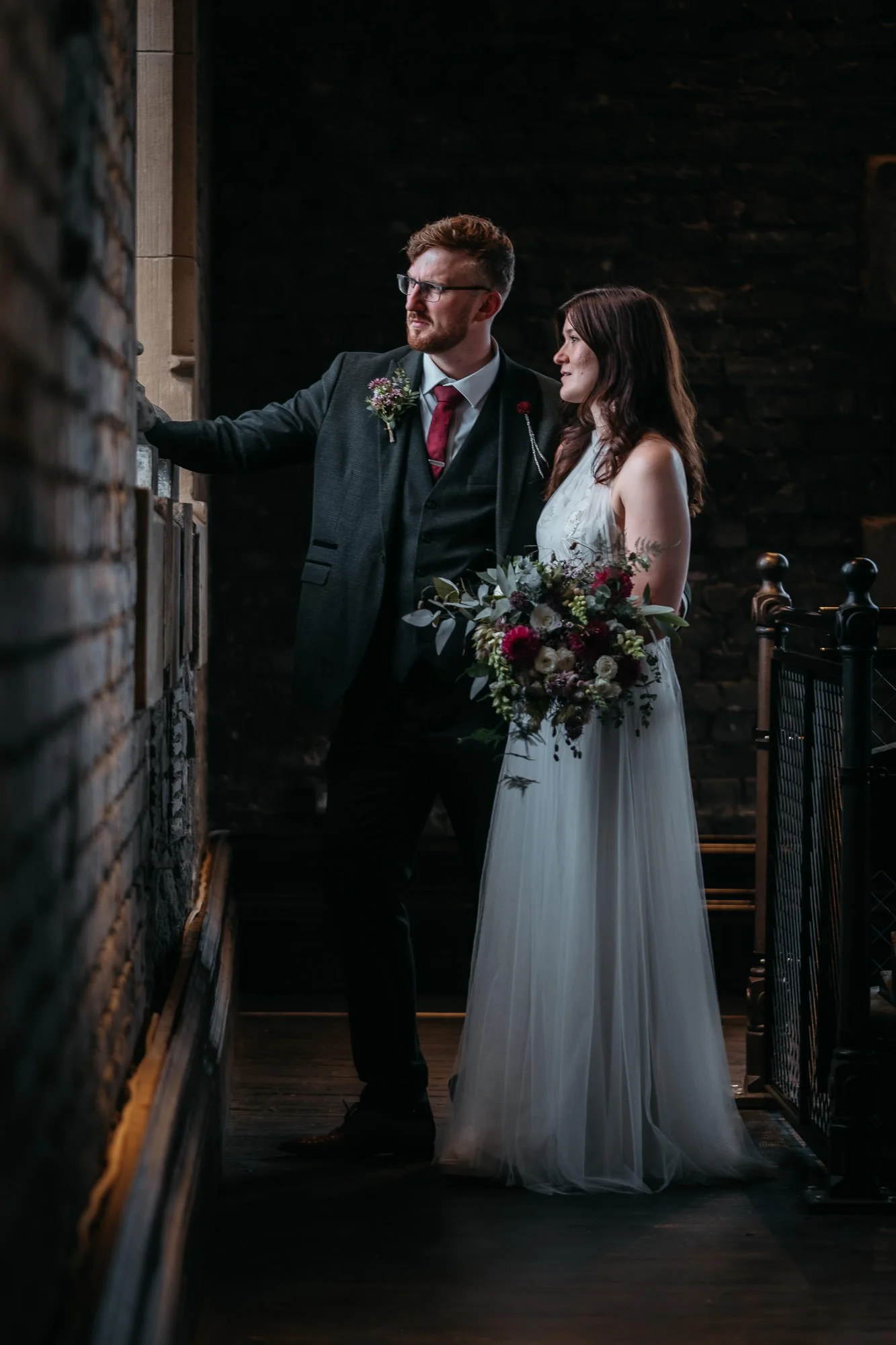 Bride and groom looking at display, groom in dark suit with boutonniere, bride in white gown holding bouquet, dimly lit interior with brick and wood walls.