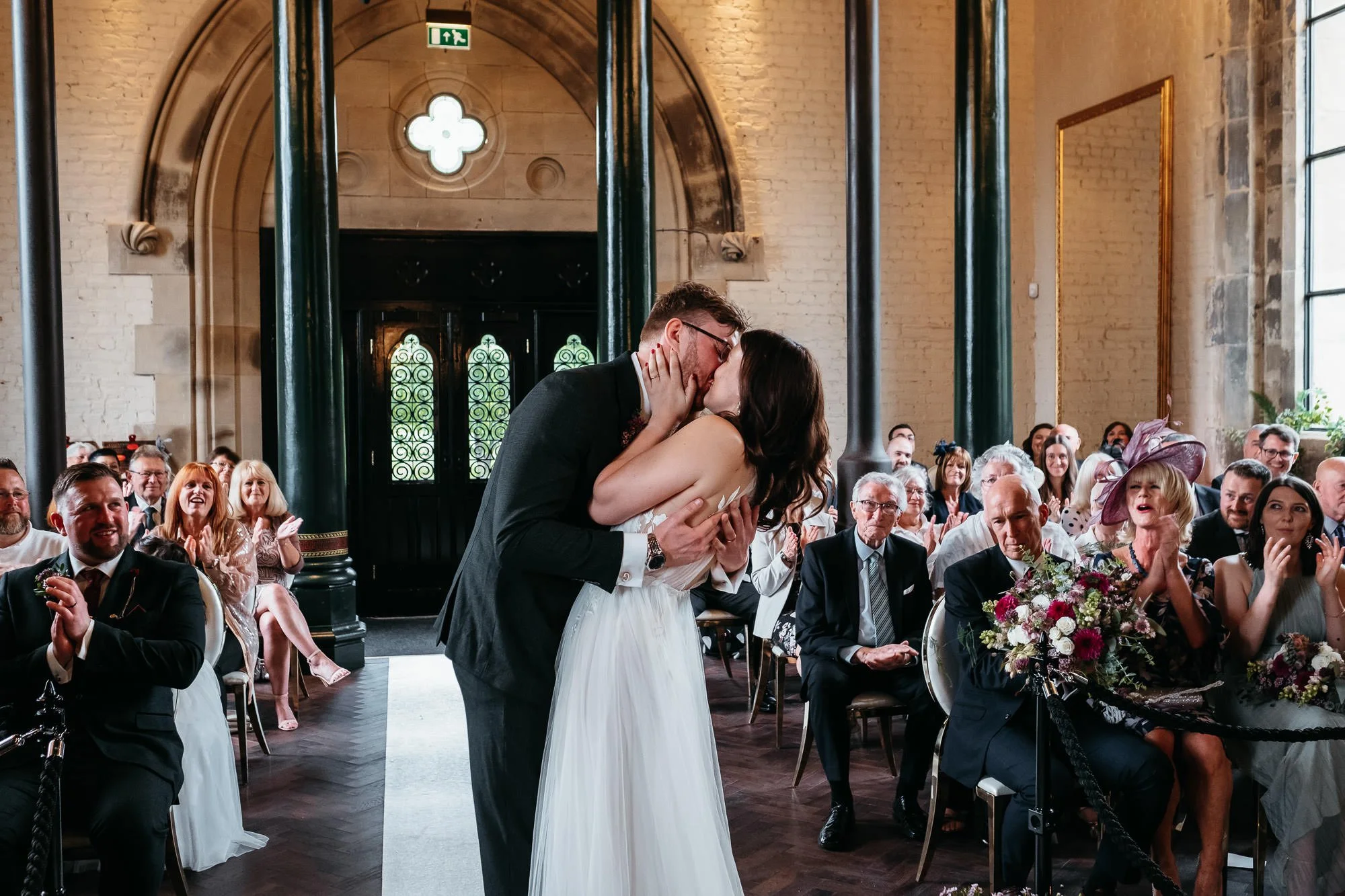 A bride and groom kiss during their wedding ceremony, surrounded by seated guests clapping and smiling in a church with high arched windows and brick walls.