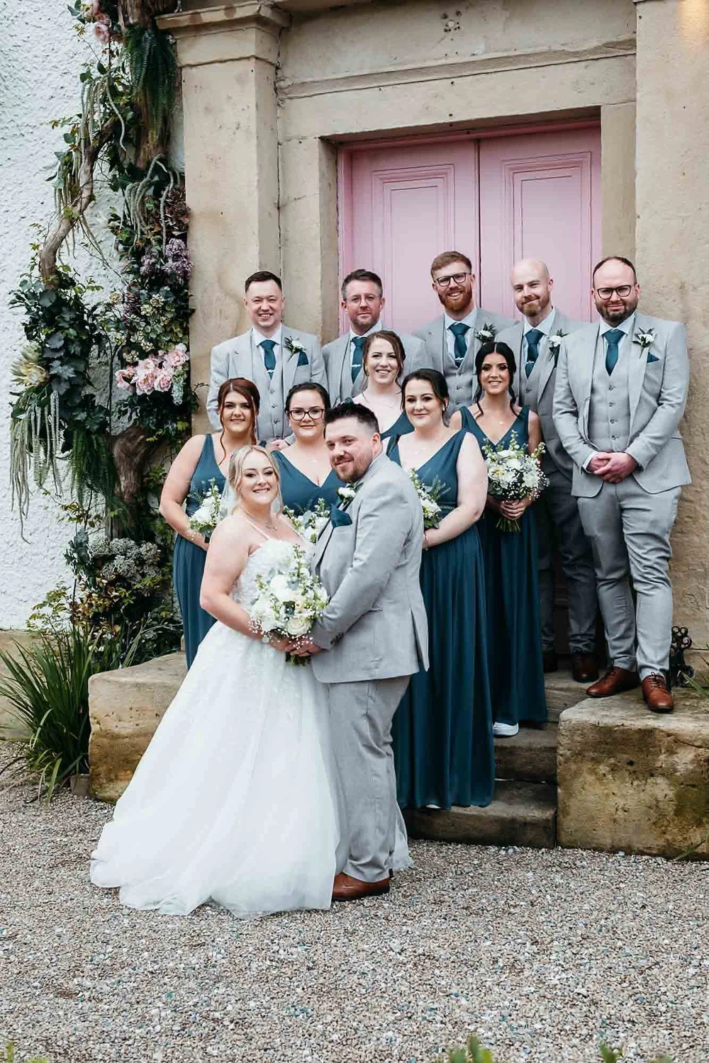 A wedding party of eleven in grey suits and teal dresses on the steps at the pink door of The Croft Hotel