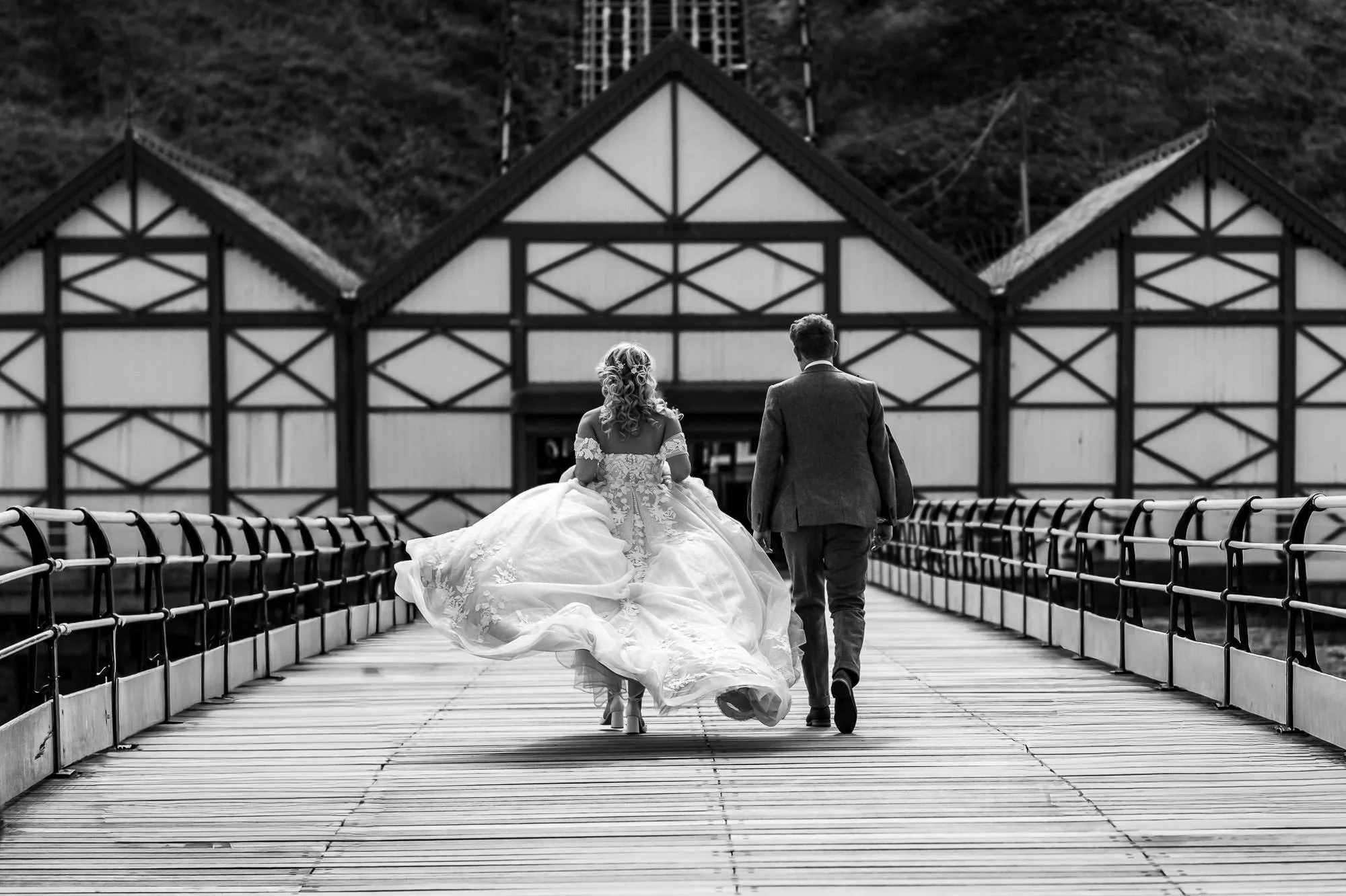 A bride in a wedding gown and a groom in a suit walking away on a bridge, with a building in the background.