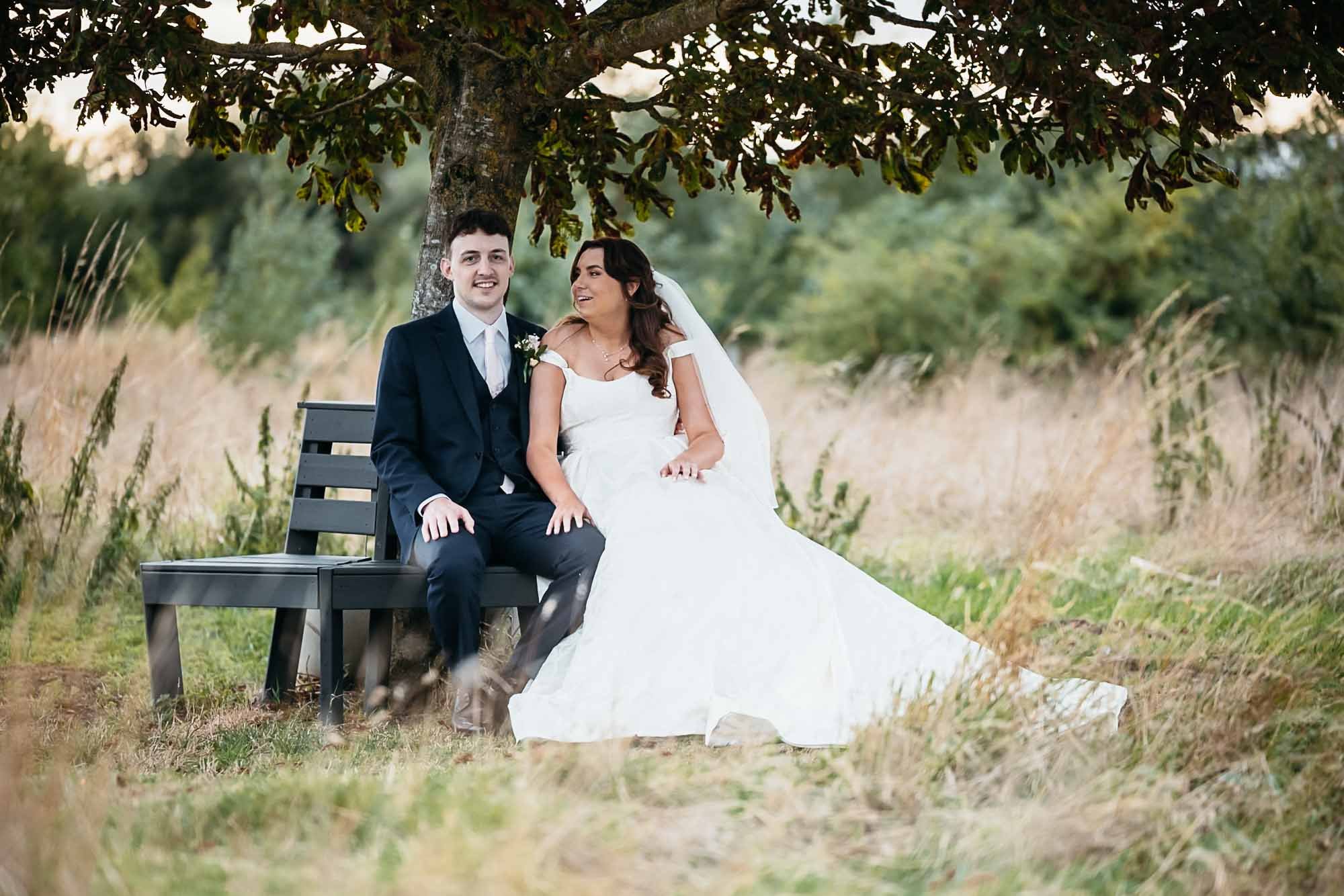 Aimee and Dan sitting together on a bench under a tree in the grounds