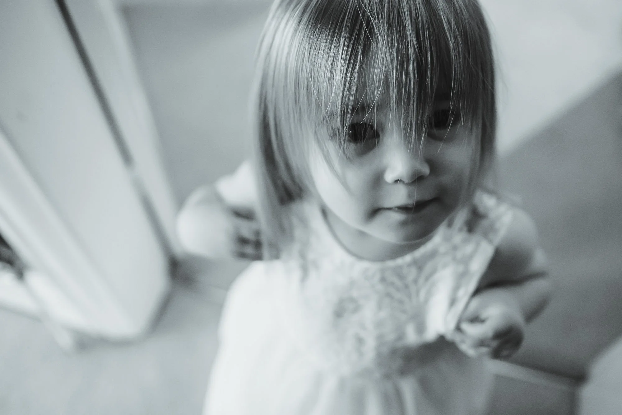 A young girl with short hair wearing a lace dress, looking up at the camera with a slight smile, indoors near a door.