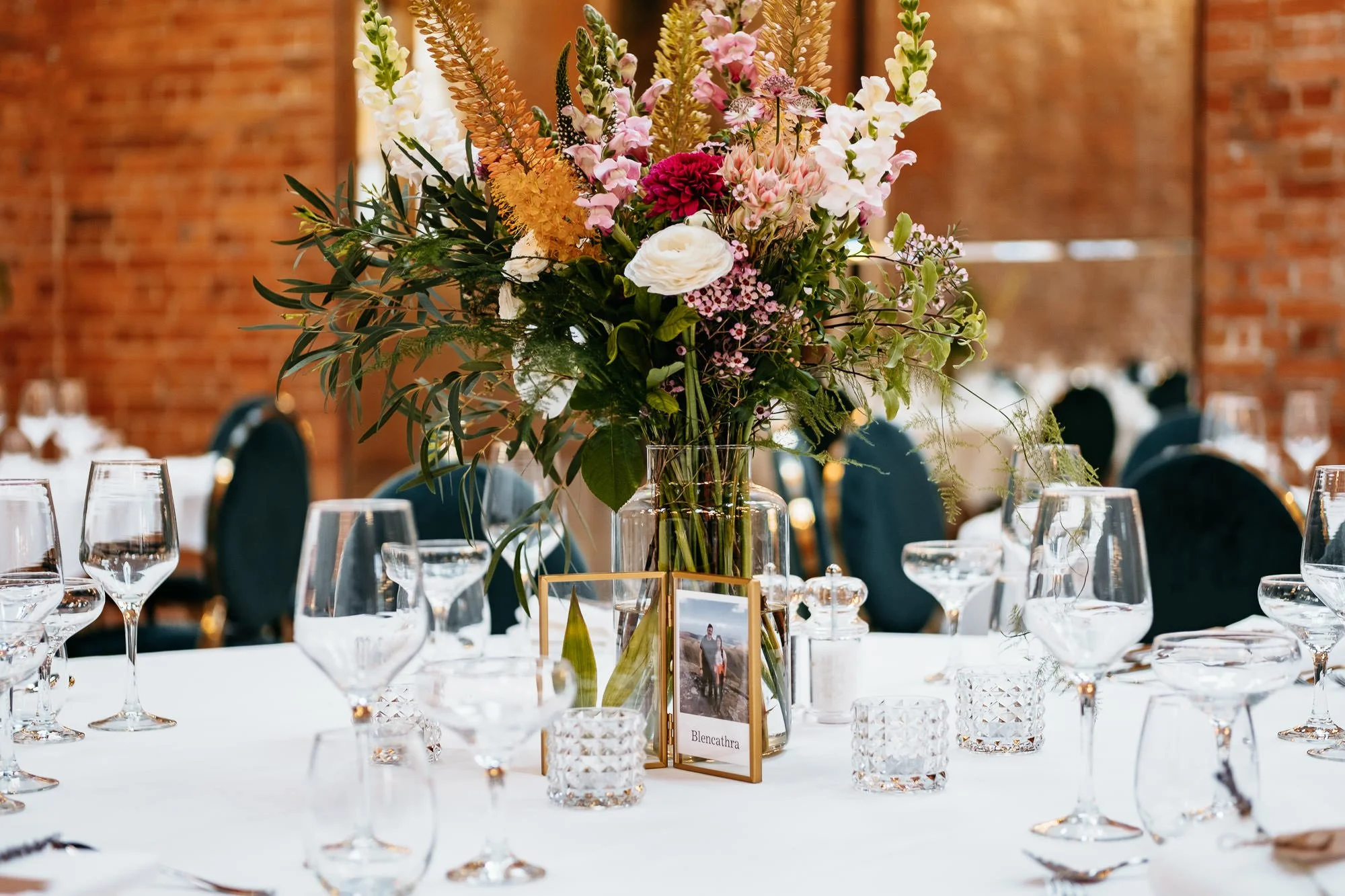A wedding reception table setting with a tall floral centerpiece in a glass vase, surrounded by multiple wine glasses, candles, and framed photos, all on a white tablecloth with black chairs and a brick wall in the background.